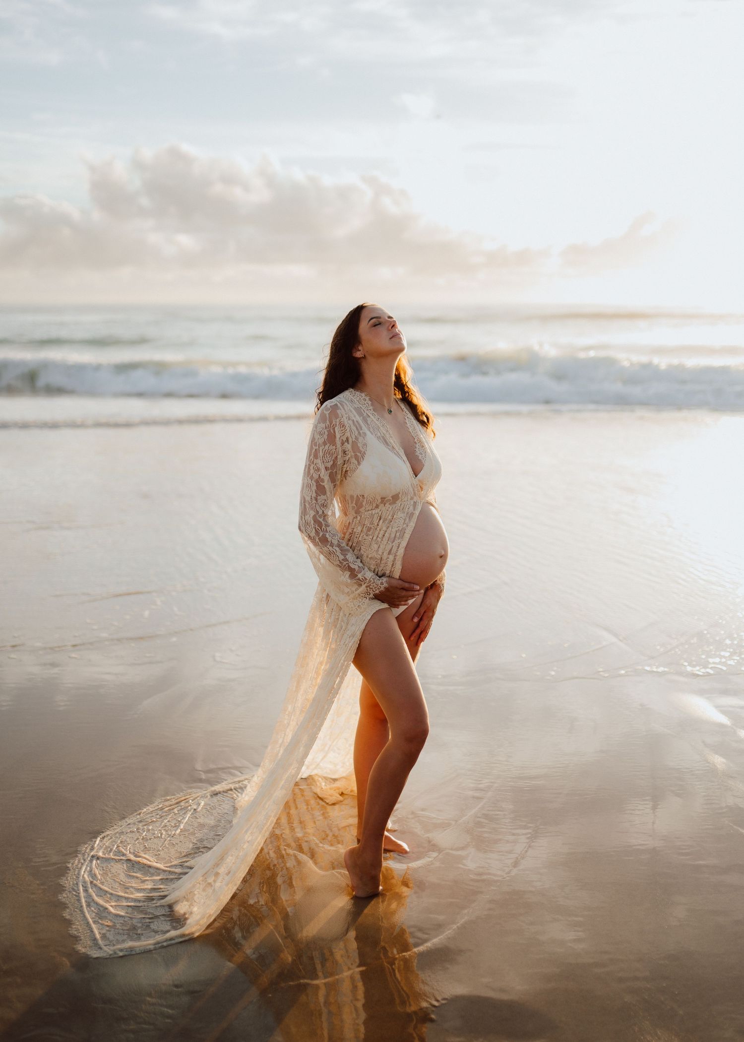 A flowing white lace dress catches the golden sunlight as waves crash on a serene beach at sunset.