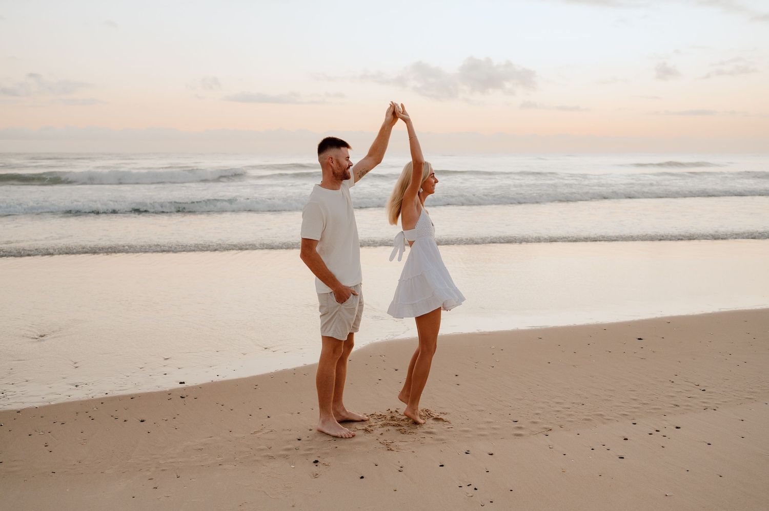 A couple in white outfits poses together romantically on a sandy beach at sunset.