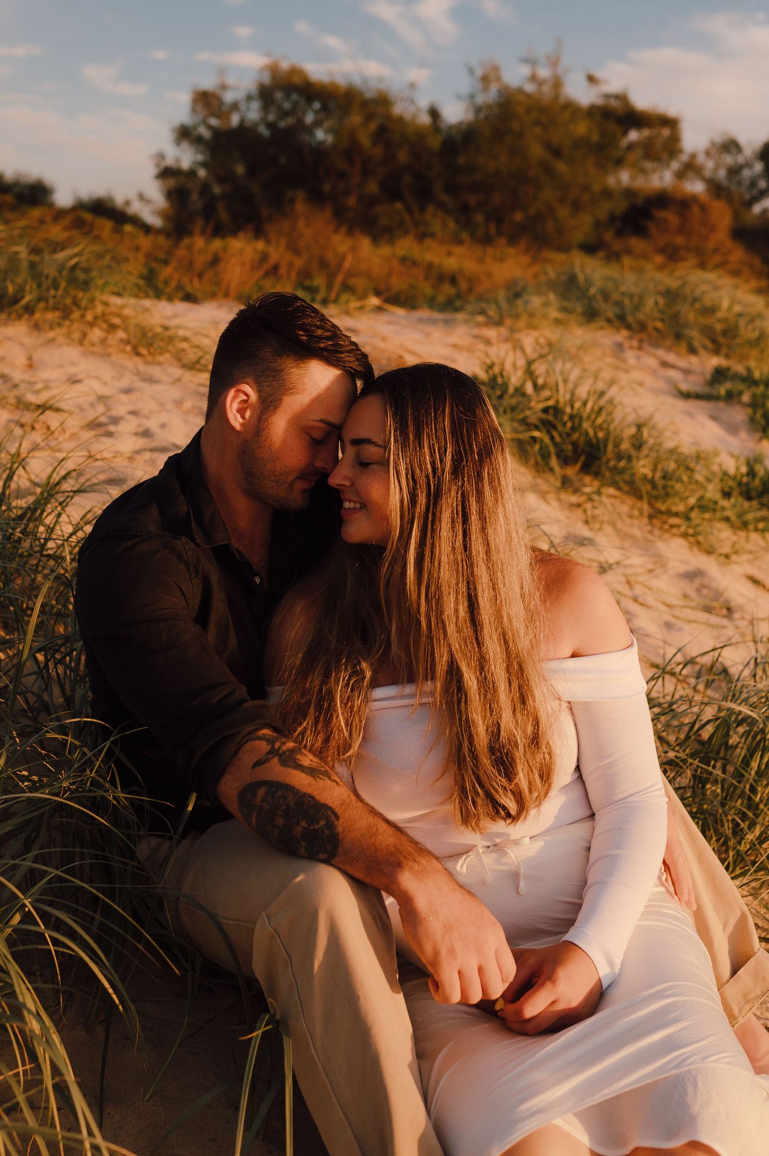 Couple sitting together in sandy dunes at golden hour, woman in white off-shoulder dress, man with tattoos embracing her.