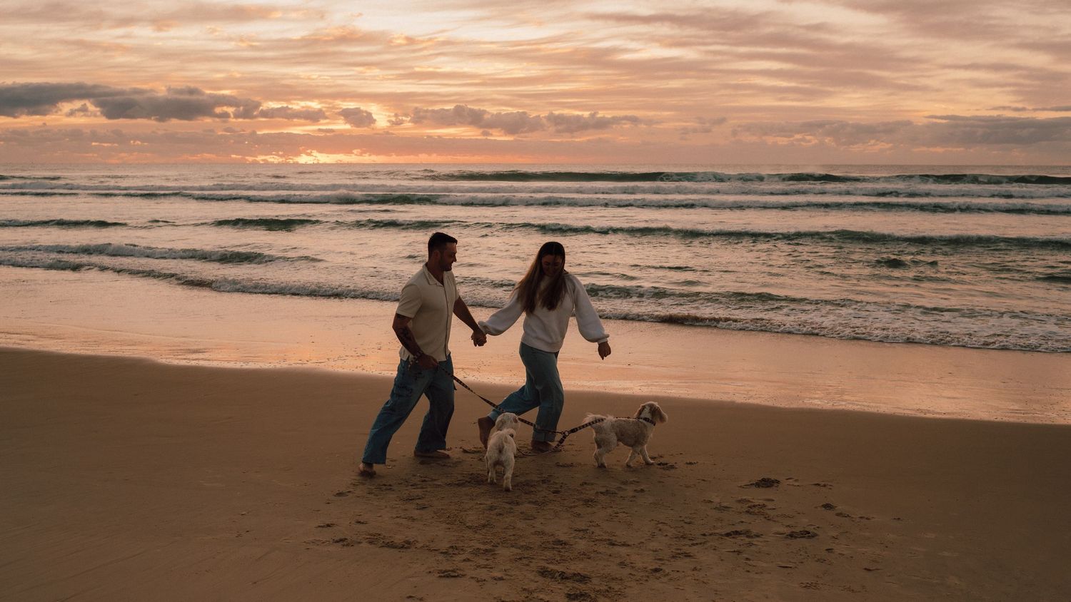 Couple holding hands walking dog on sandy beach at sunset with dramatic cloudy sky and ocean waves.