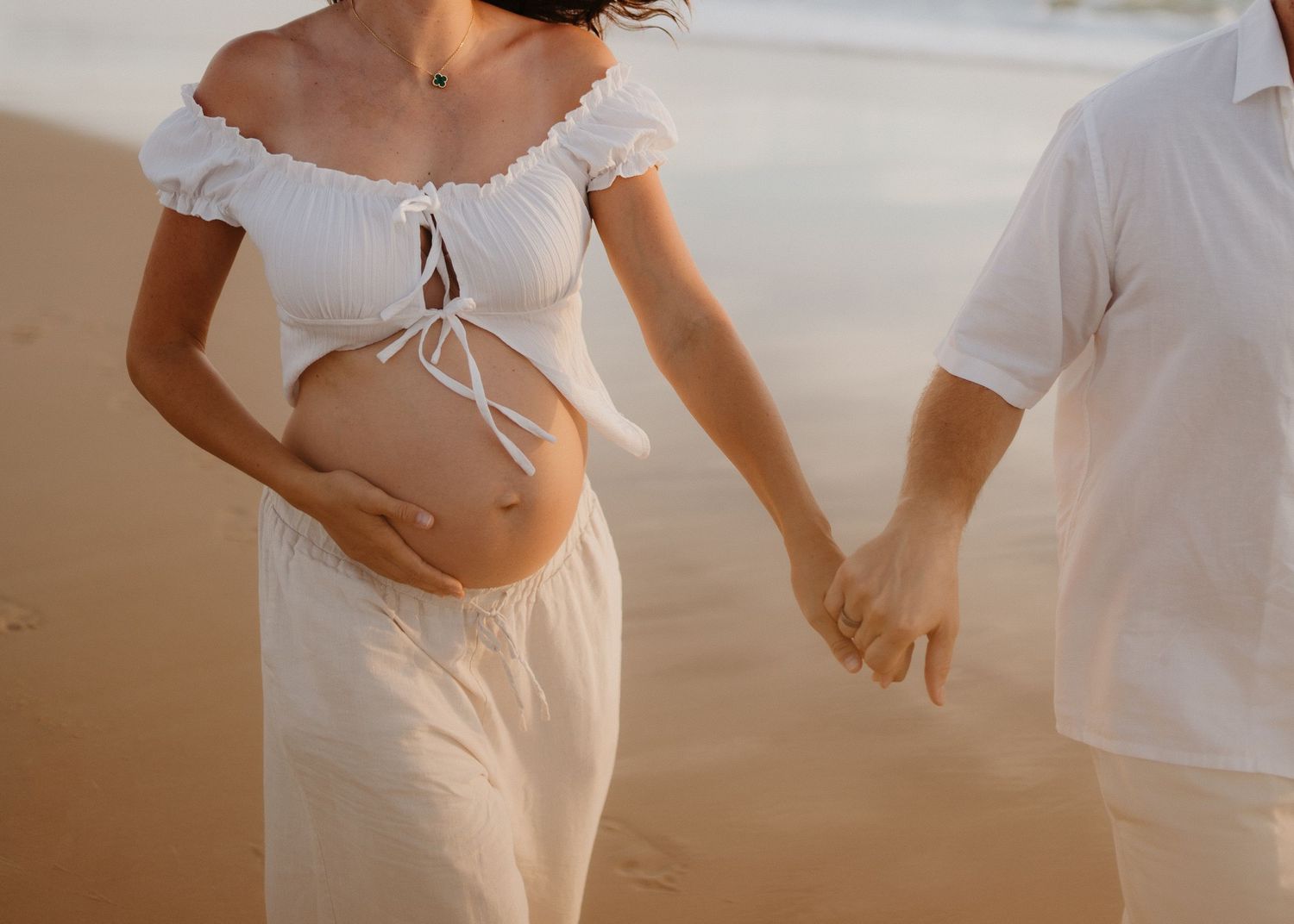 Couple holding hands on beach with pregnant woman in white dress showing bare belly during maternity photoshoot.