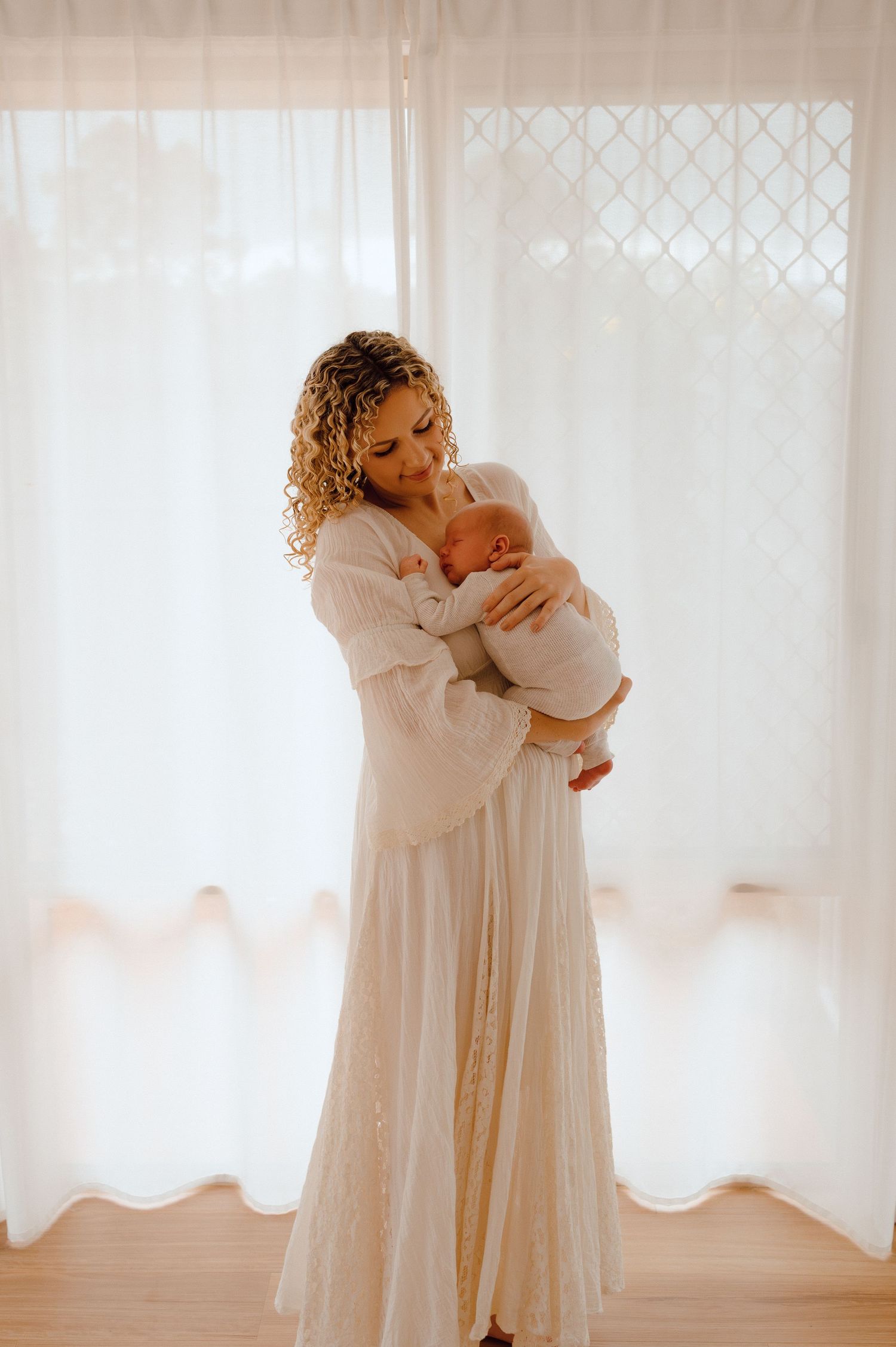 Curly-haired woman in white dress tenderly holds newborn baby near sheer curtained window in soft natural light.