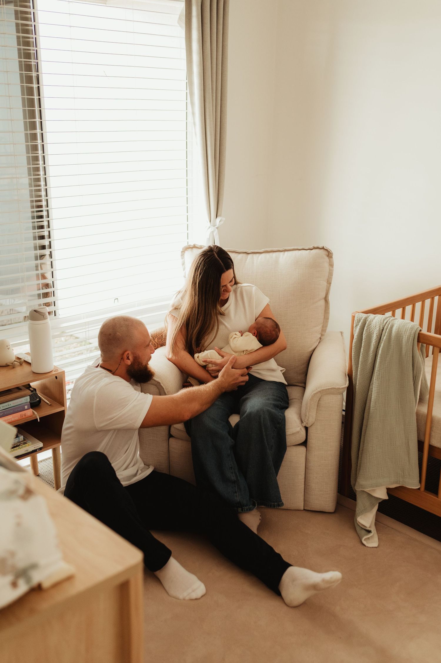 New parents sitting together in nursery, mother holding newborn baby in armchair while father sits on floor beside her.