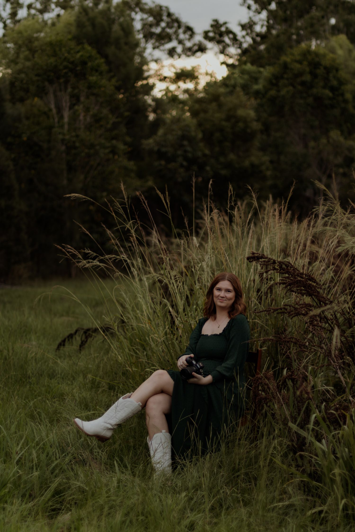 Person in a dark green dress poses gracefully in tall grass during a golden hour outdoor photoshoot.