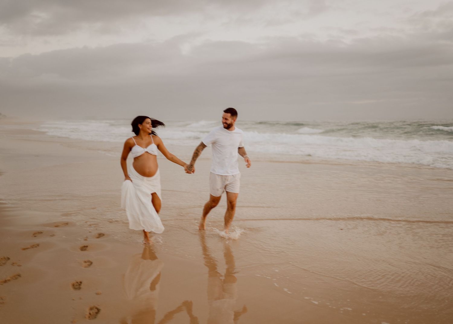 A couple in white clothing walks together on a sandy beach leaving footprints during sunset.