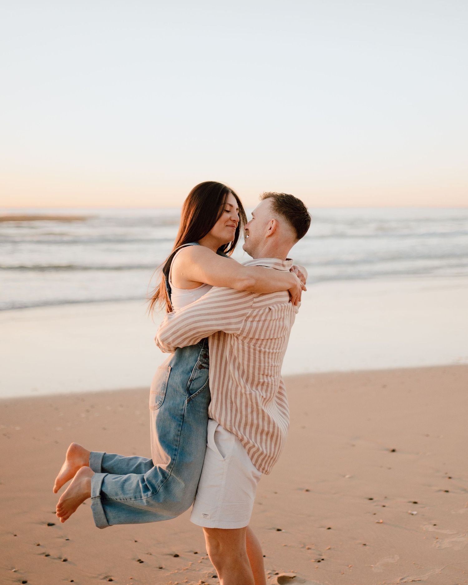 A couple shares a playful moment lifting each other on a beach during golden hour.