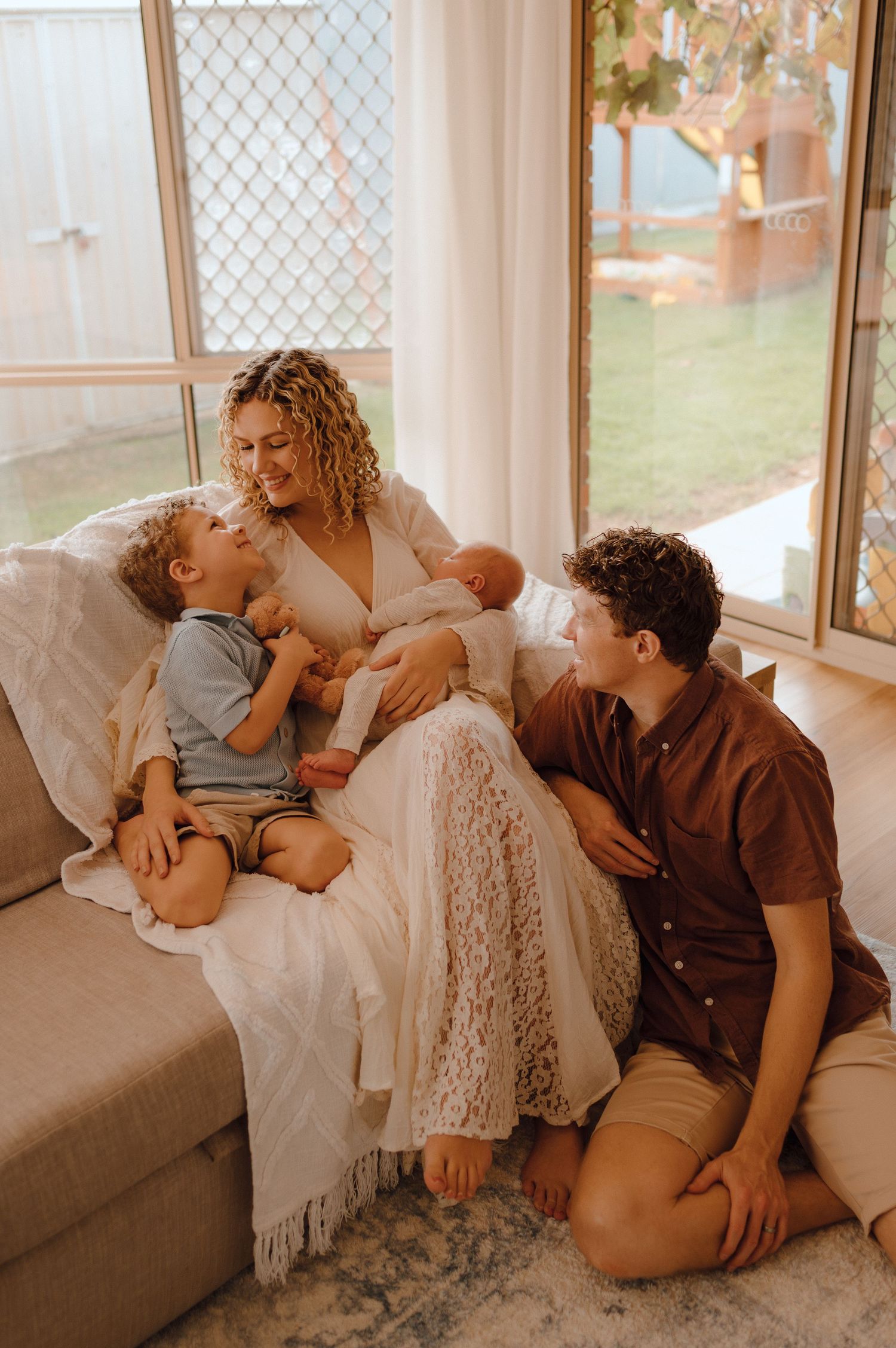 A joyful family with a newborn baby relaxes together on a couch near bright windows in a cozy living room.