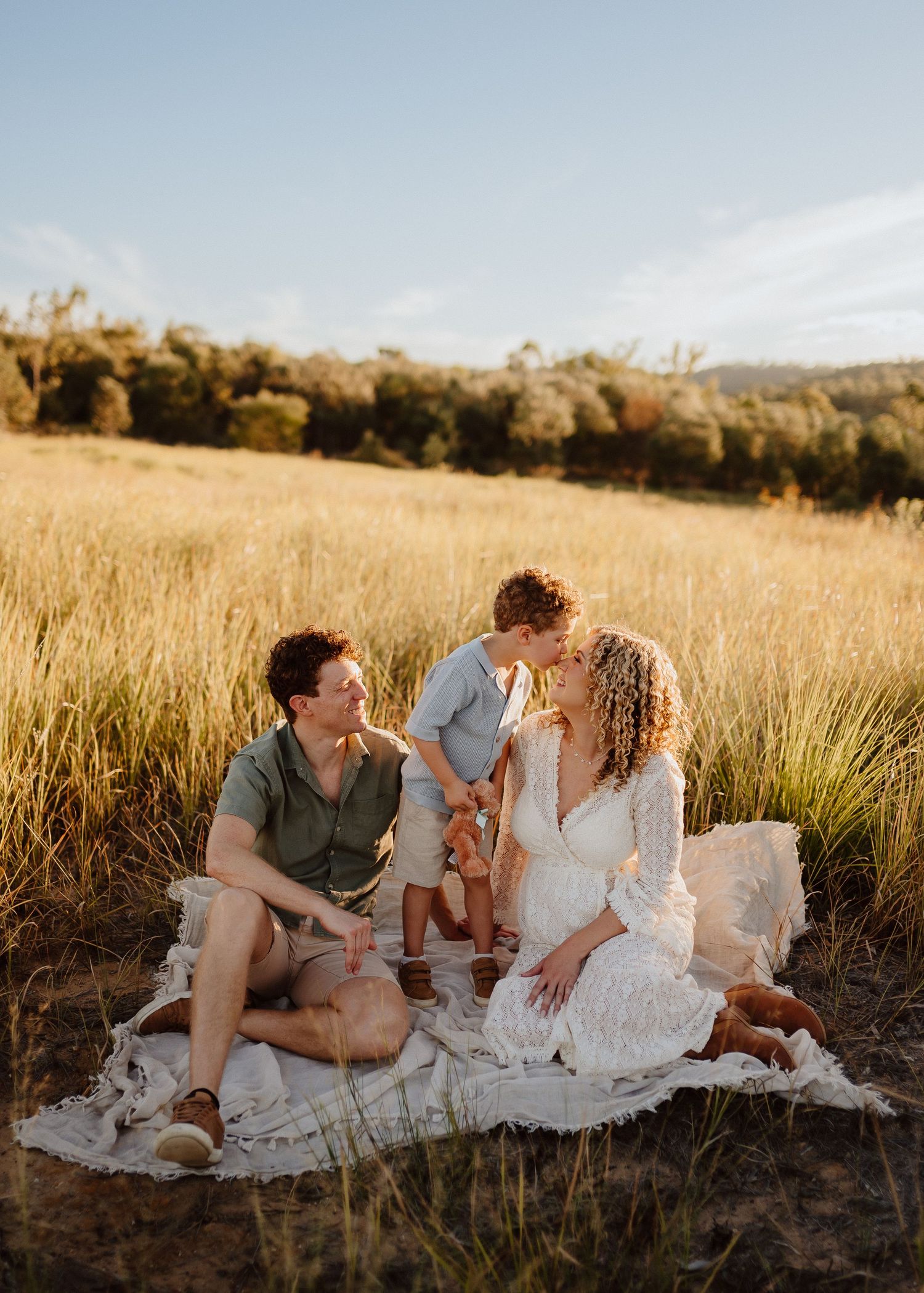 A family enjoys a sunset picnic on a blanket in a golden wheat field with mountains in the background.
