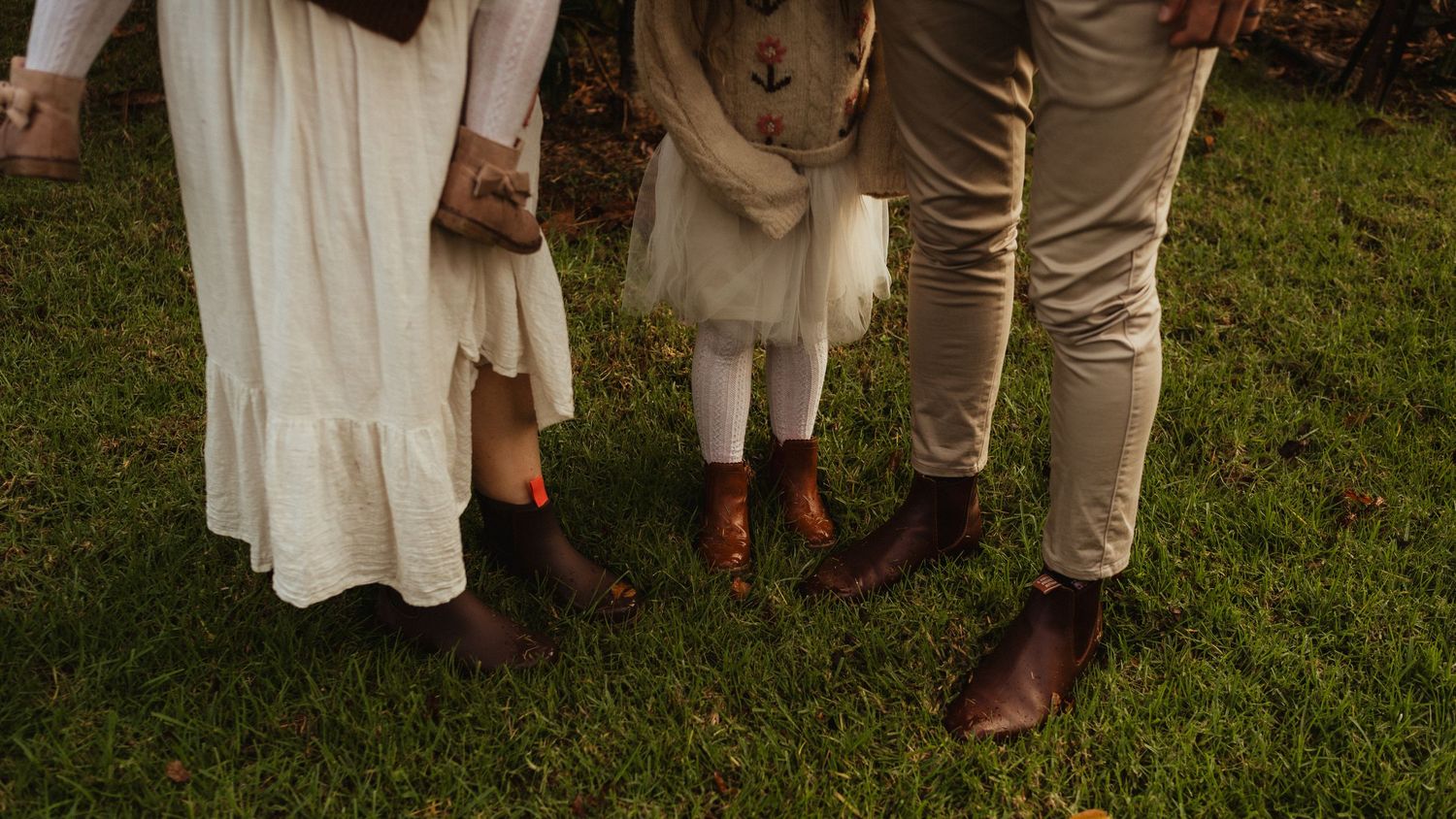 Family of three standing on grass, wearing matching brown Chelsea boots, white dress, beige pants, and a childs white outfit.