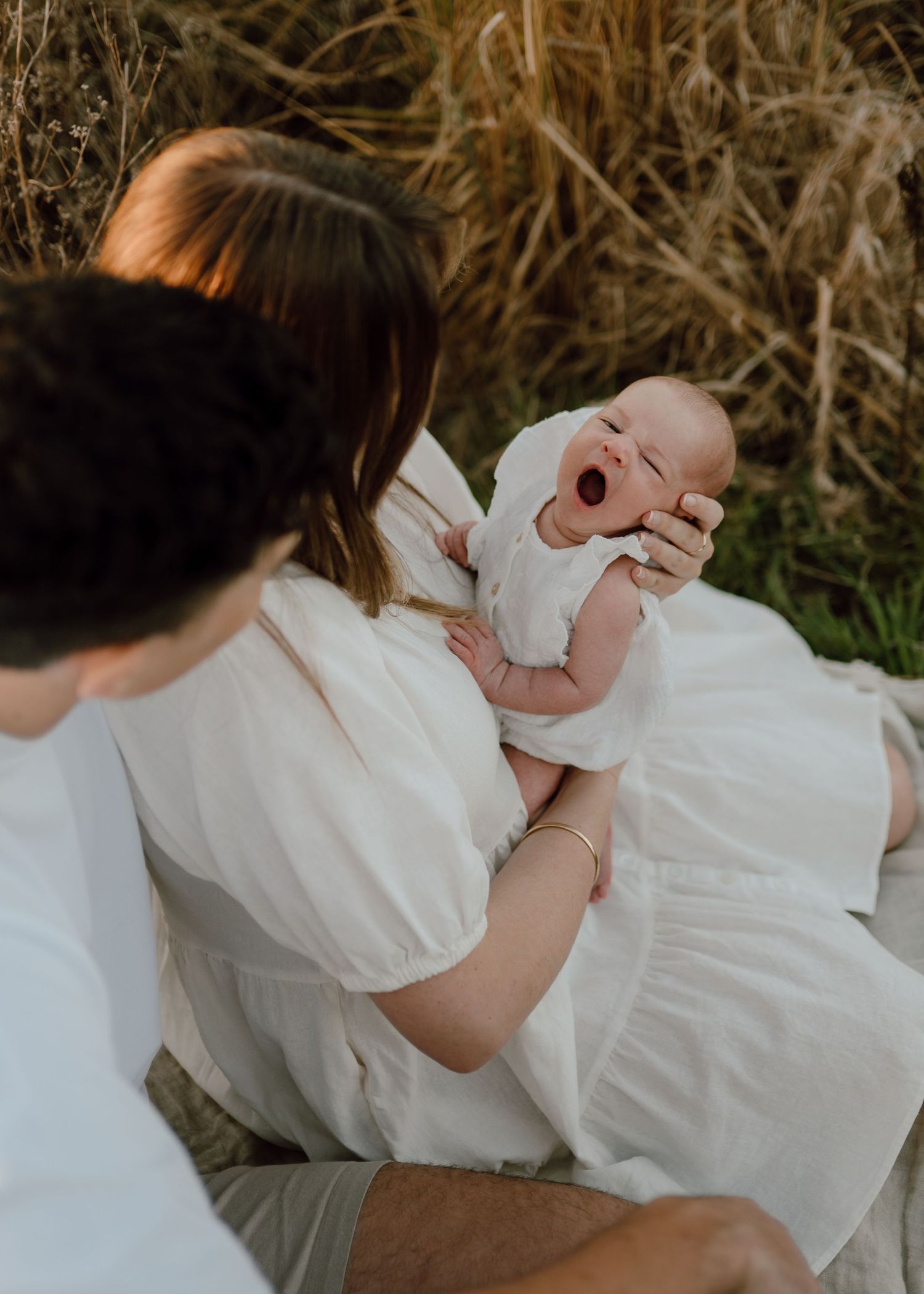 A baby in a white dress lies on grass during an outdoor photoshoot.