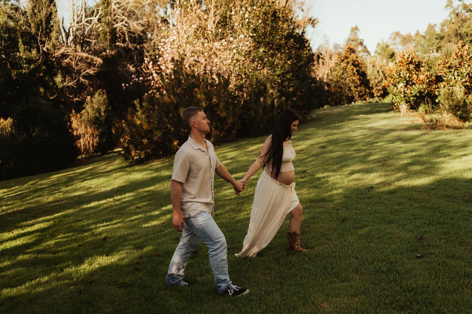 Couple holding hands walking across green grass lawn surrounded by lush trees in warm golden sunlight.