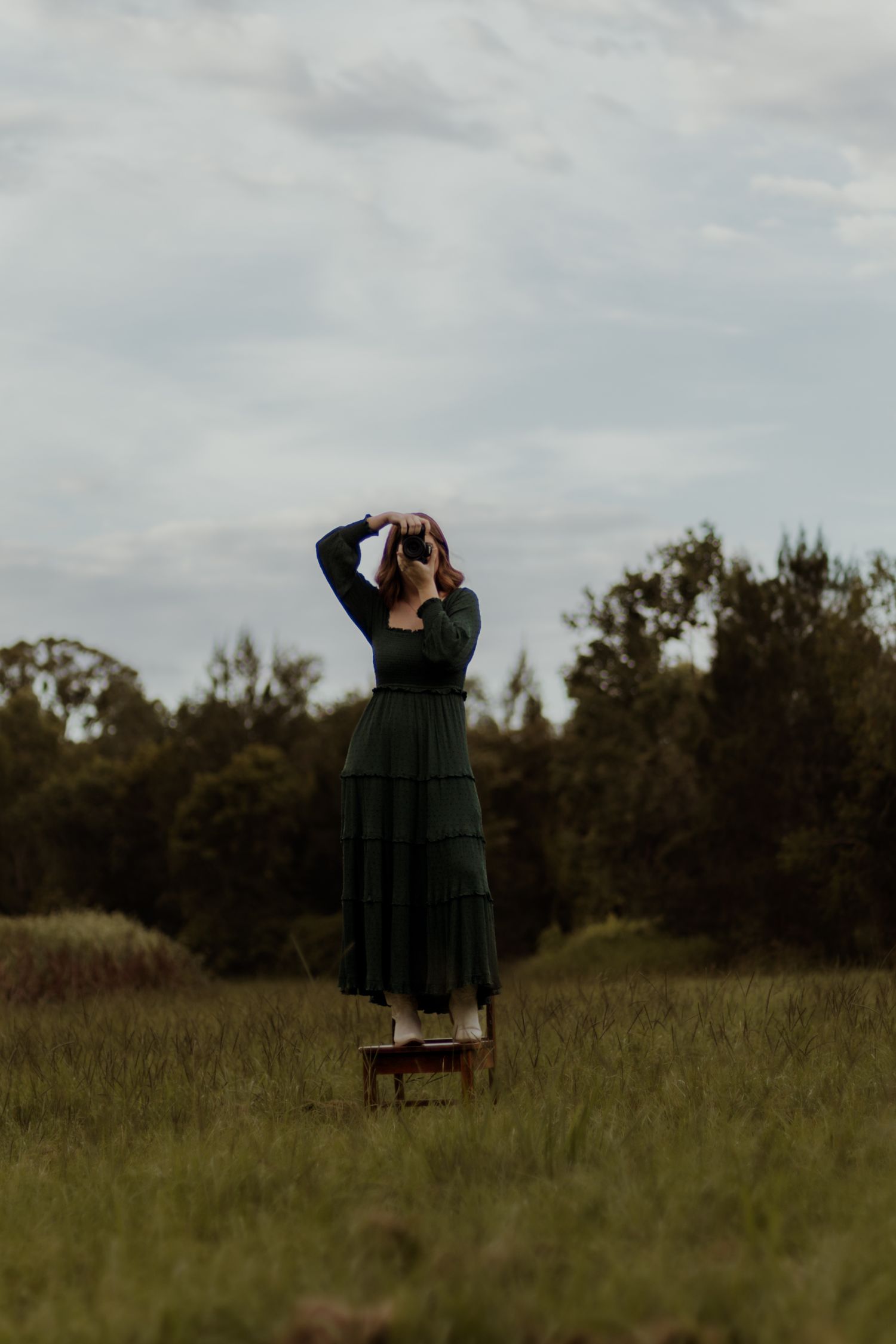 A figure in a dark green vintage dress stands on a wooden stool in a misty field at dusk.