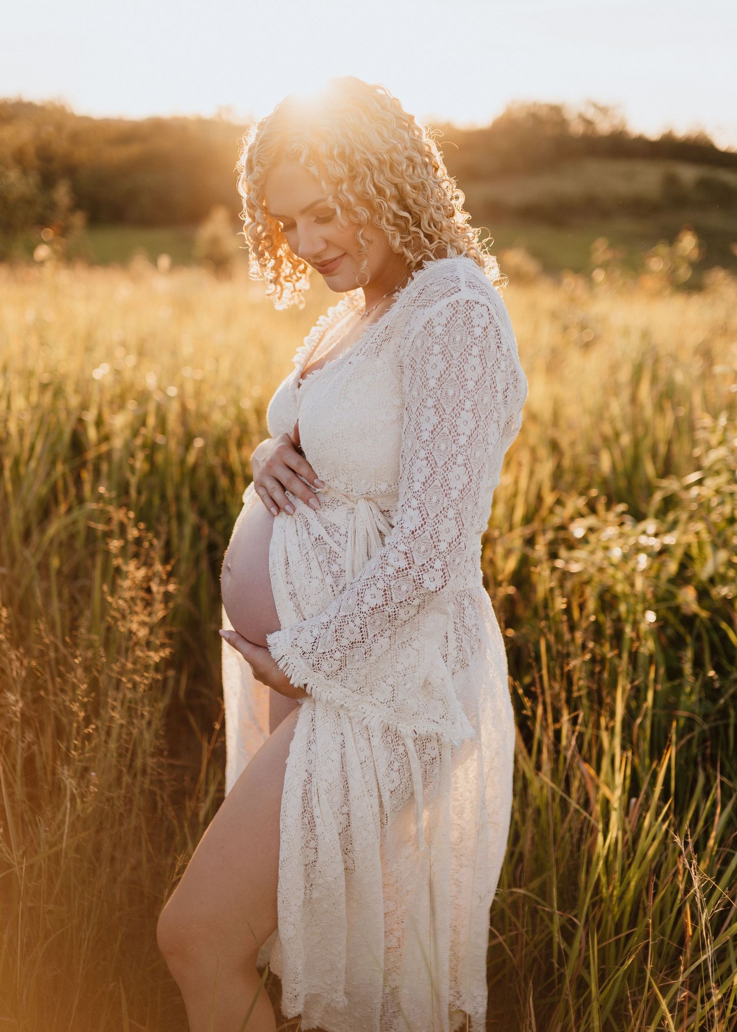 A dreamy maternity photo in a golden field at sunset shows an ethereal white lace robe and flowing curls backlit by warm light.