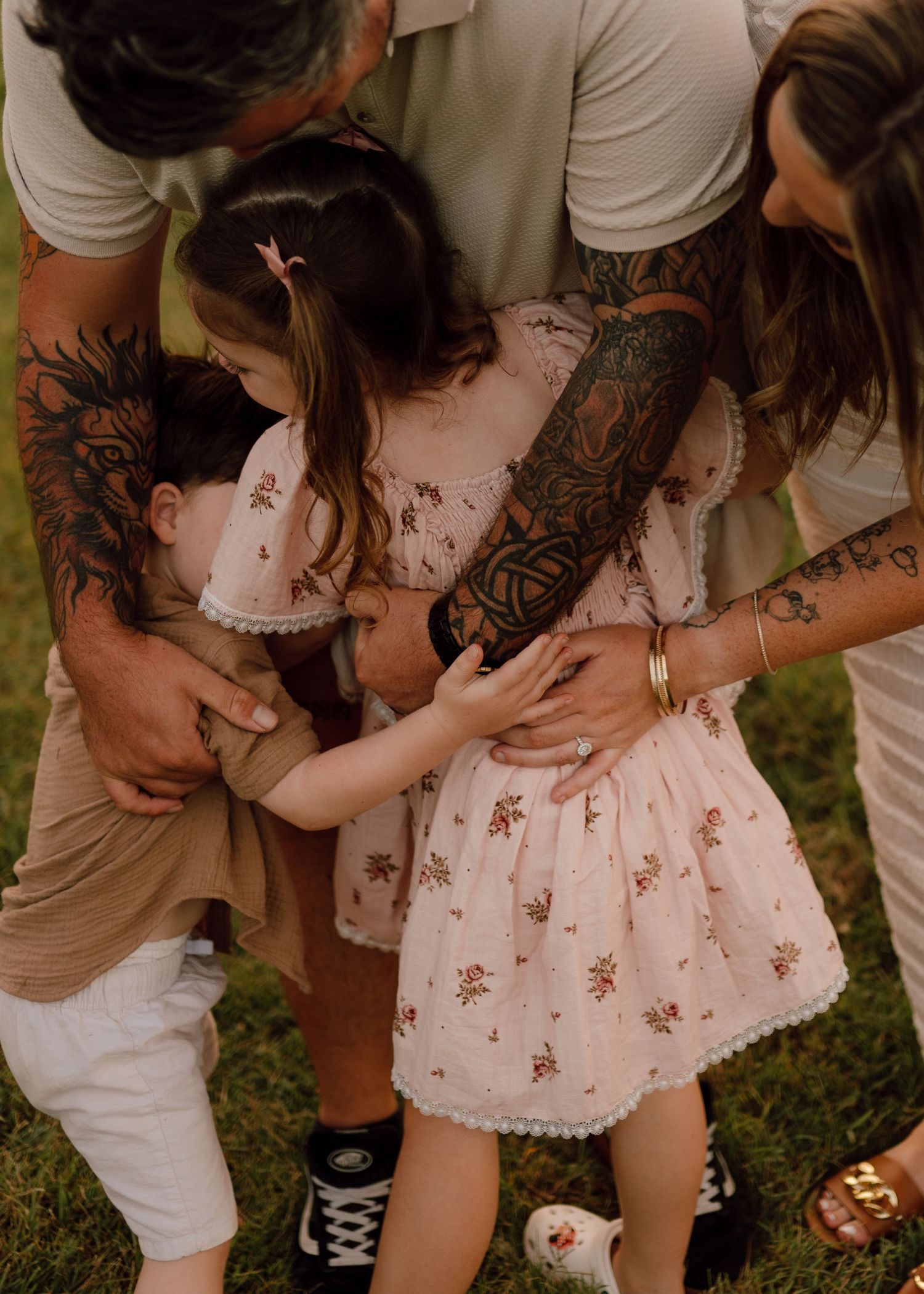 A family wearing white and beige clothing embraces outdoors, showing off their artistic tattoo sleeves.