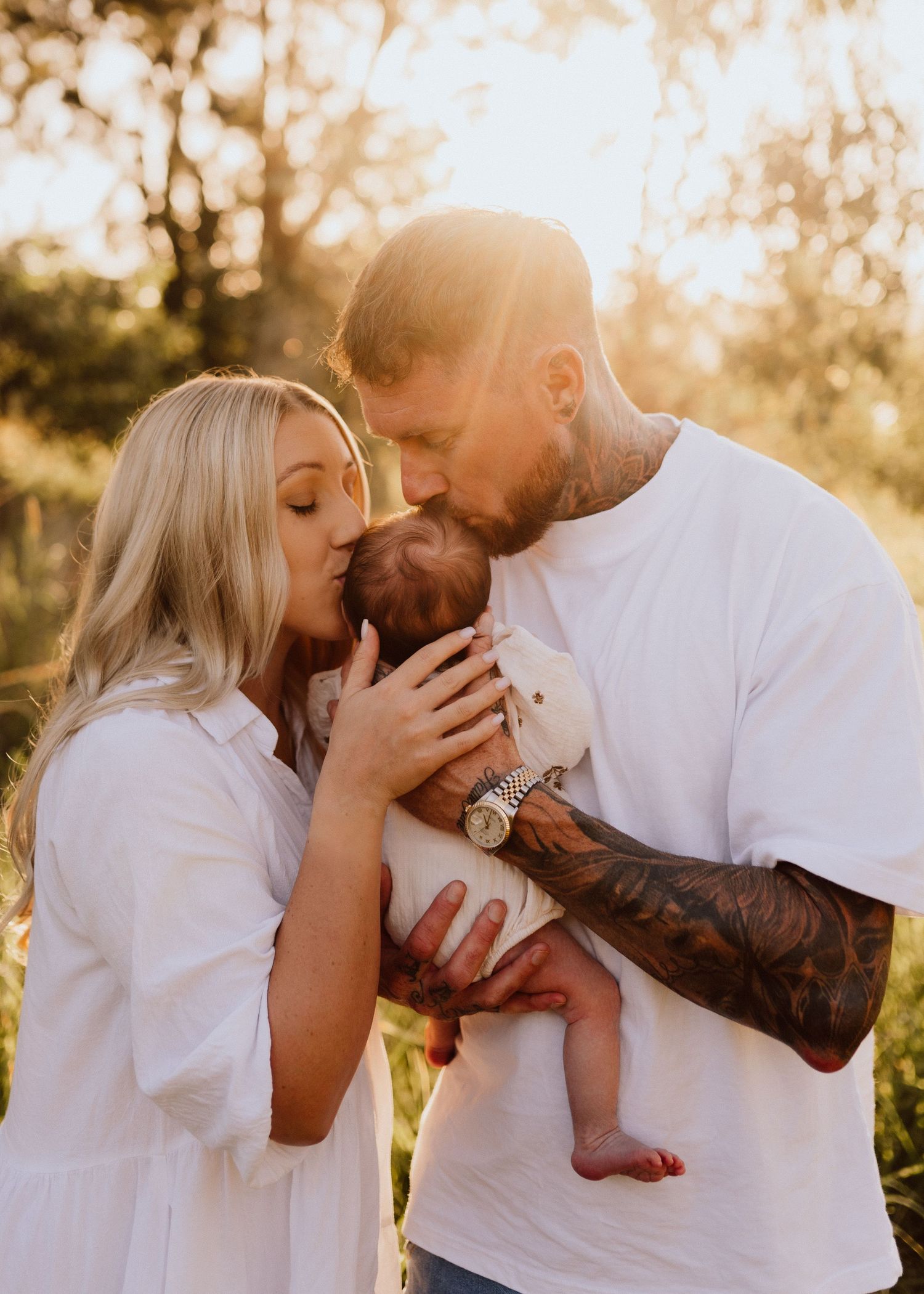 A tattooed couple in white outfits kisses their newborn baby outdoors in golden sunlight.