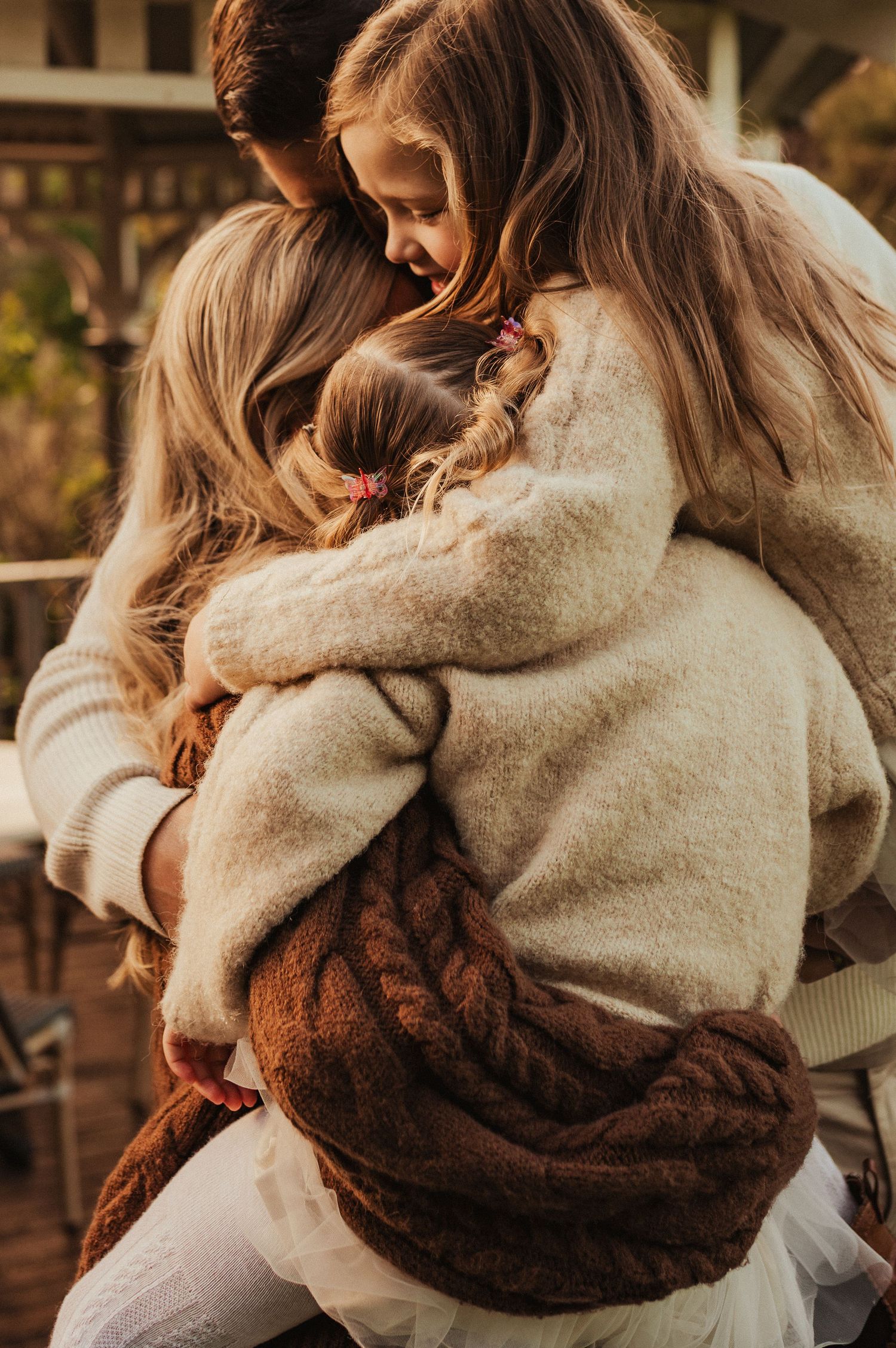 A mother tenderly embraces her two young daughters in warm cozy sweaters outdoors in soft golden light.