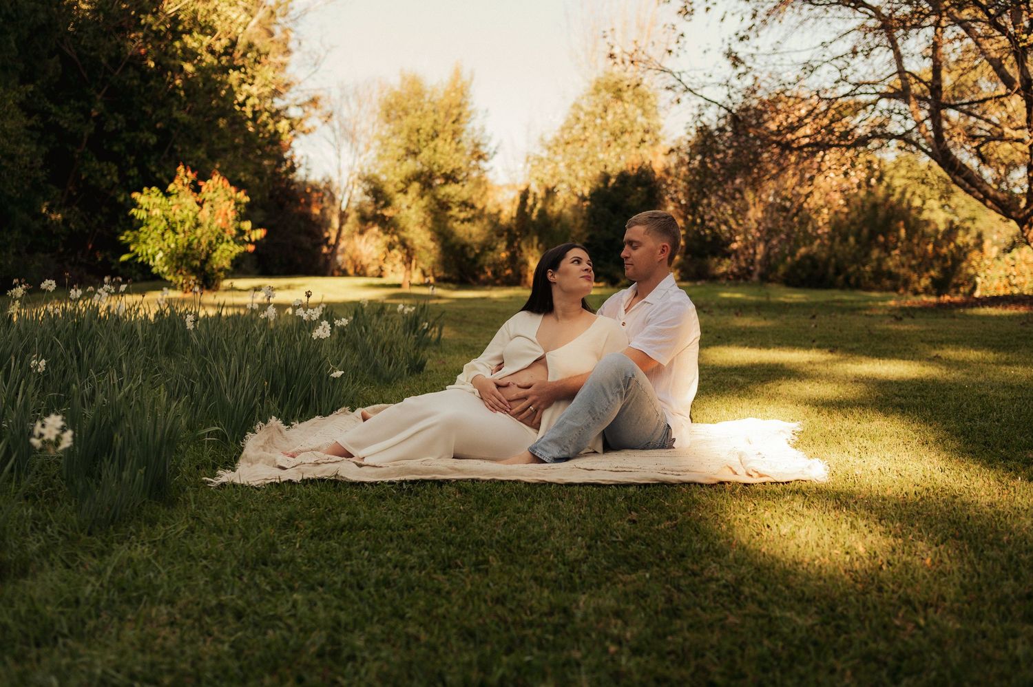 Couple sitting on a blanket in a sunny garden, woman in white maternity dress leaning against partner during golden hour.