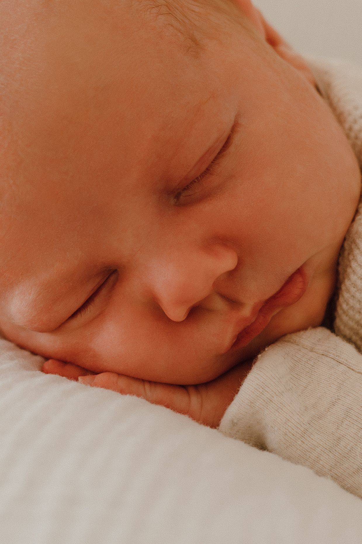 Close-up of a peacefully sleeping newborn baby resting cheek on tiny hands, wearing a cream onesie on white bedding.
