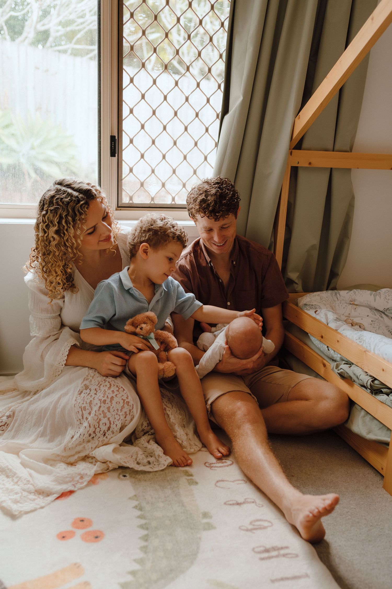 Family of four sitting on floor by window, parents smiling at newborn baby while toddler holds stuffed toy.