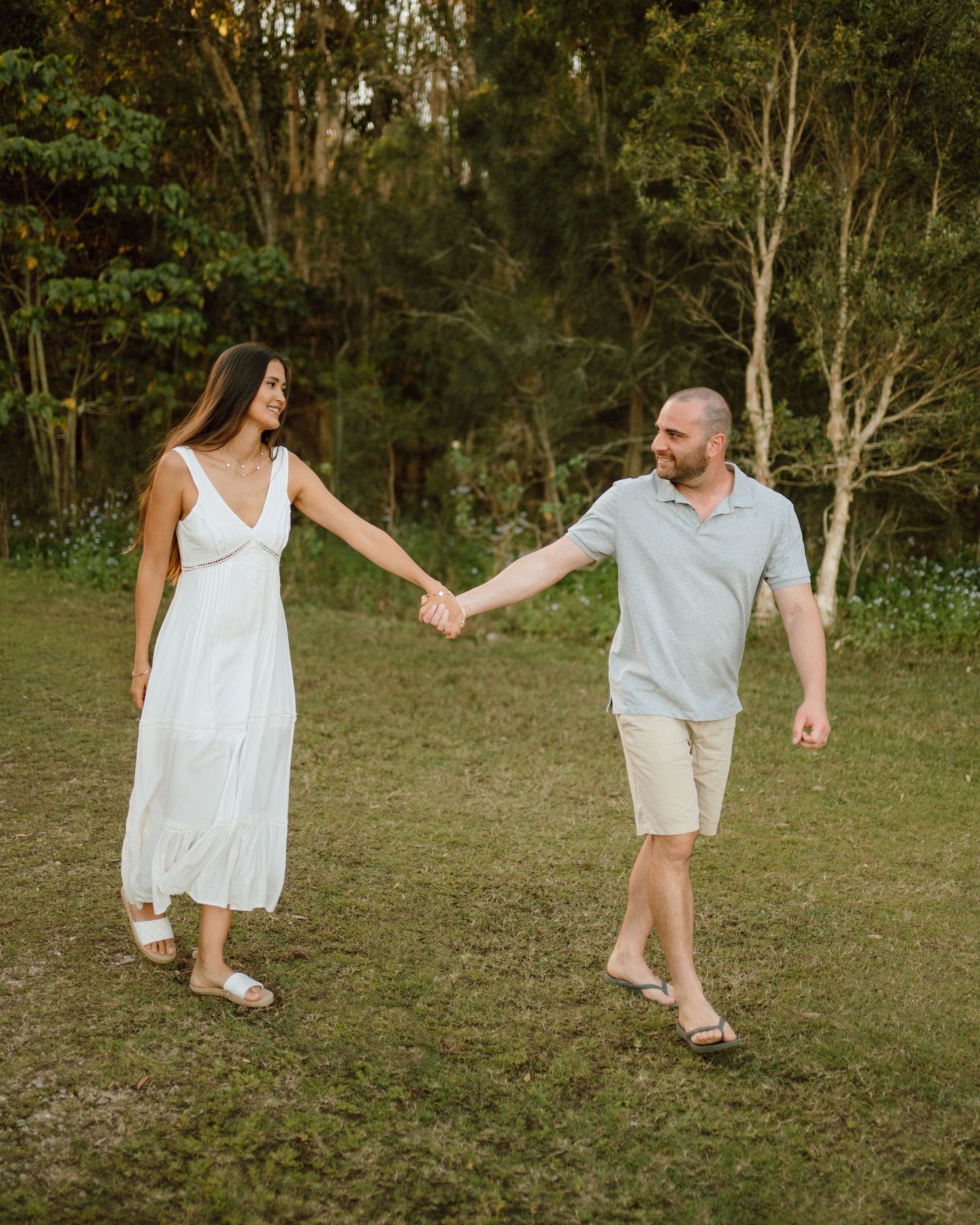 Couple walking hand in hand through a grassy field at sunset wearing white and grey casual outfits.