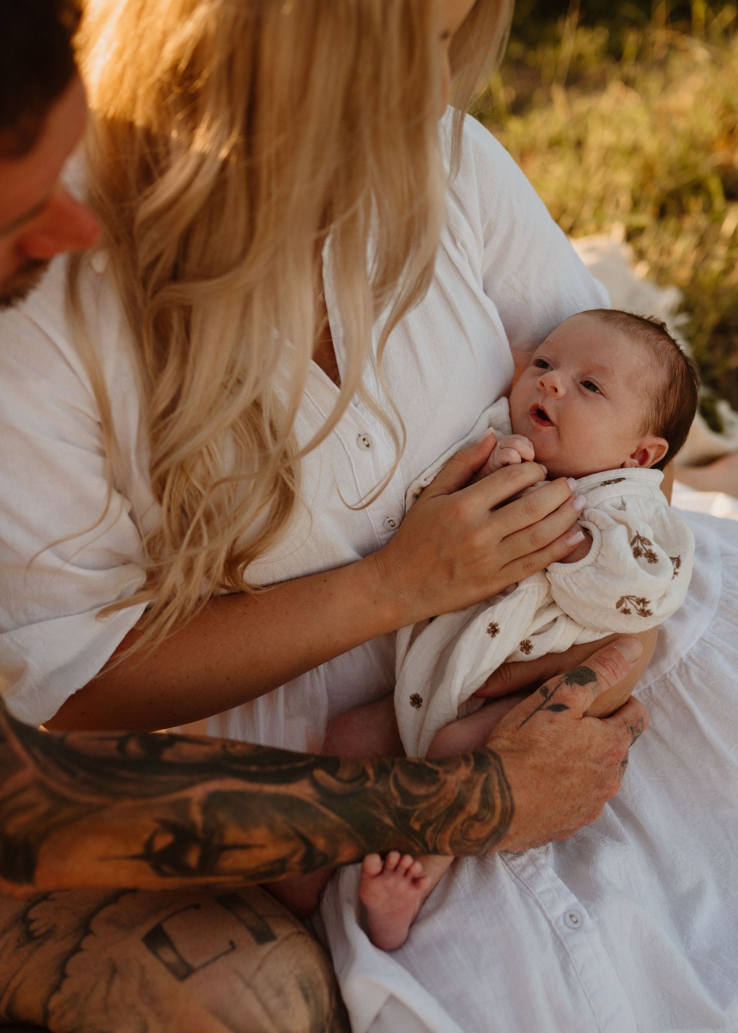 Tattooed father and blonde mother in white dress tenderly holding newborn baby outdoors.