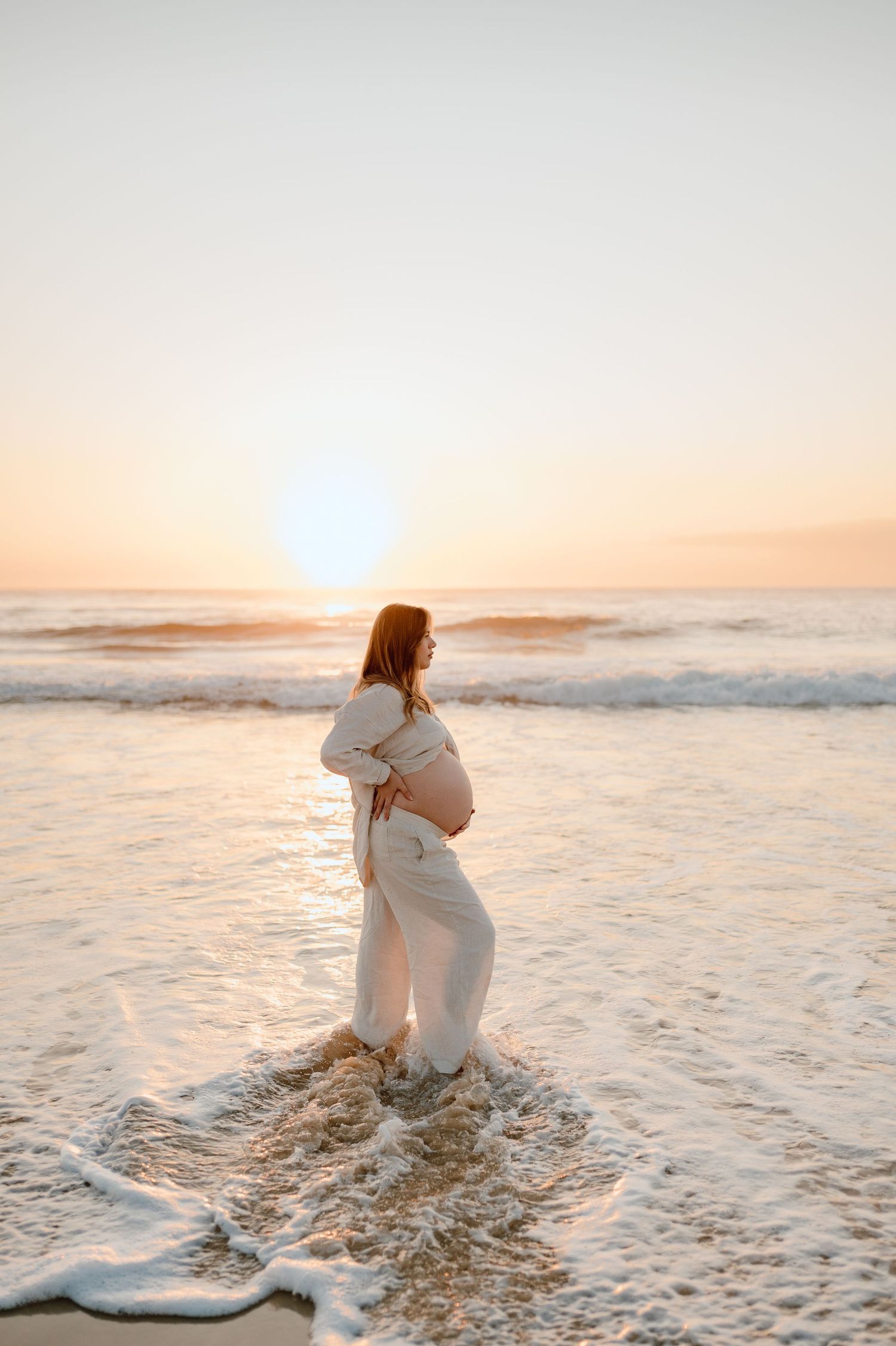 Dreamy beach maternity photos with waves crashing during sunset in white attire.