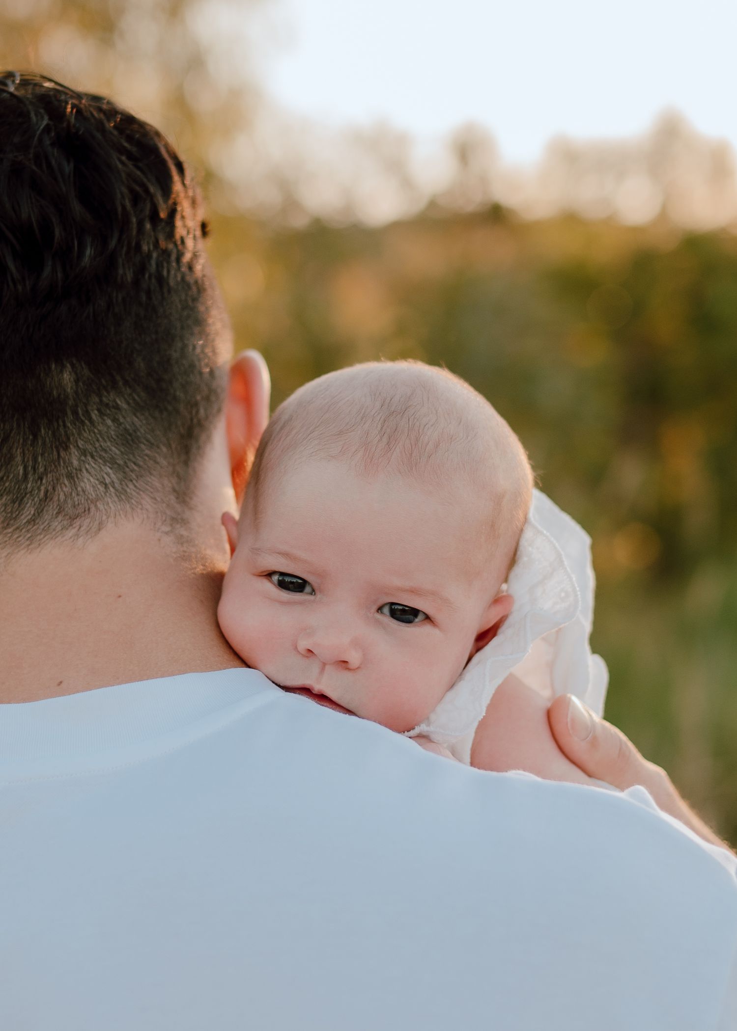 Baby looking over parent's shoulder during outdoor sunset portrait session.