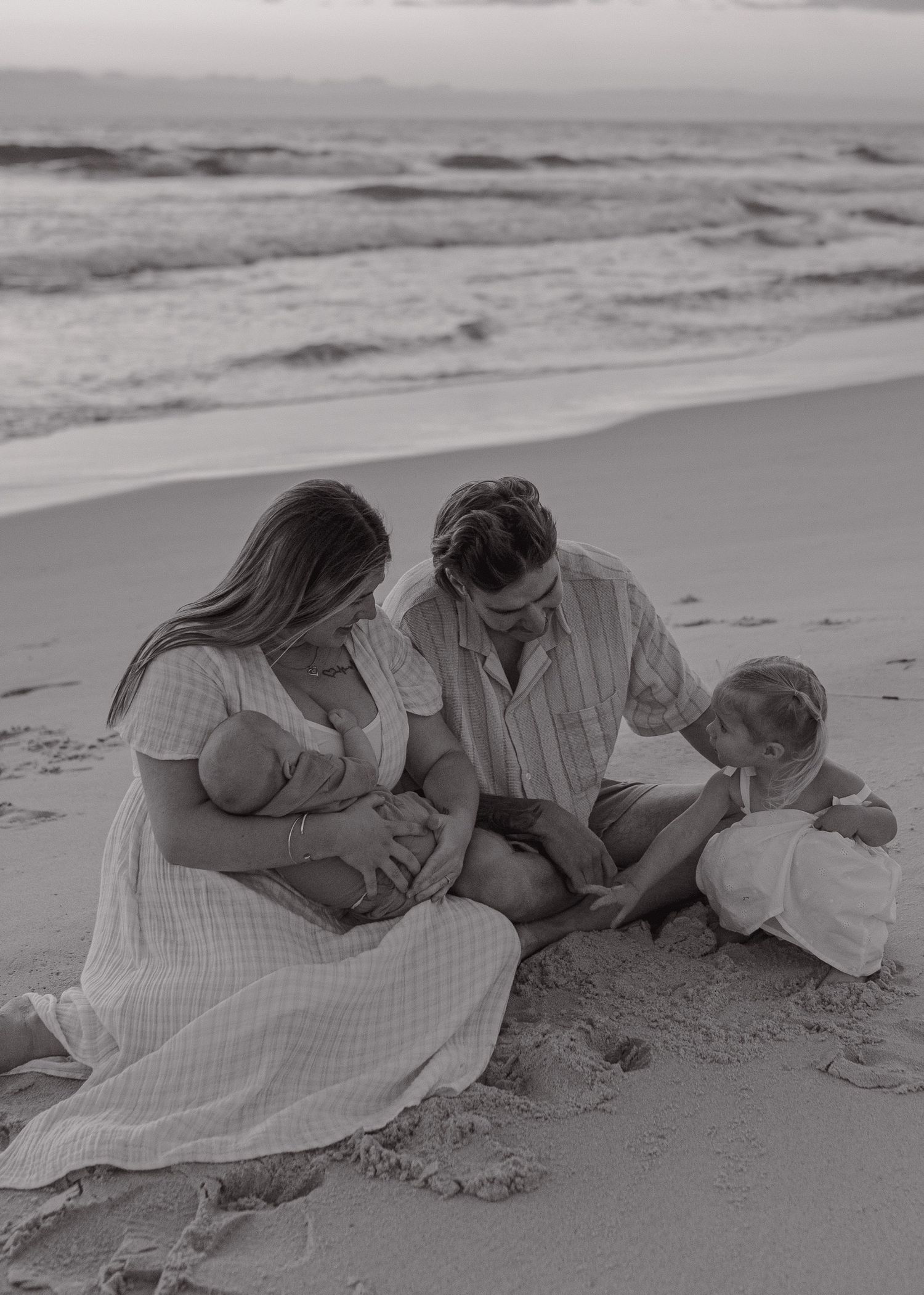 Family lying together on sandy beach at sunset in black and white.