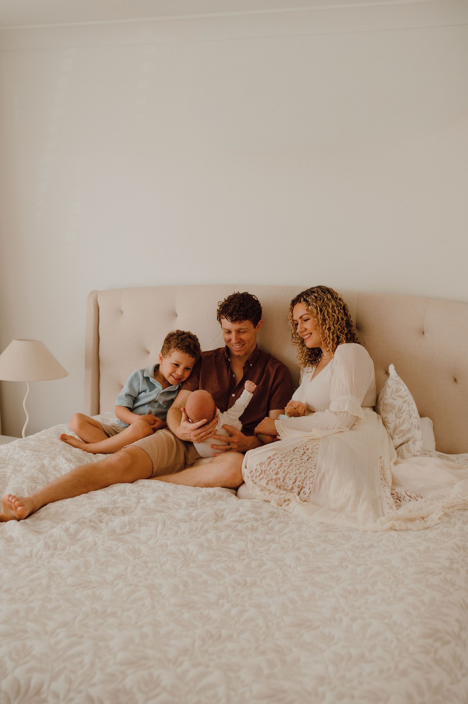 Family of four with newborn baby sitting together on a white bed, sharing a warm and tender moment.
