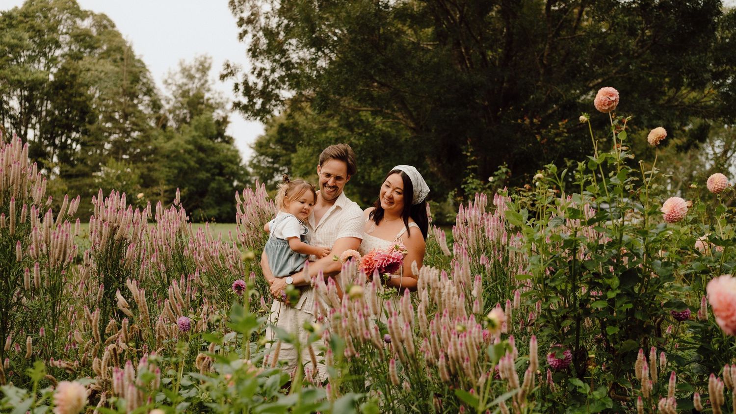 A happy family with a young child poses among blooming pink flowers and lush greenery in a beautiful garden.