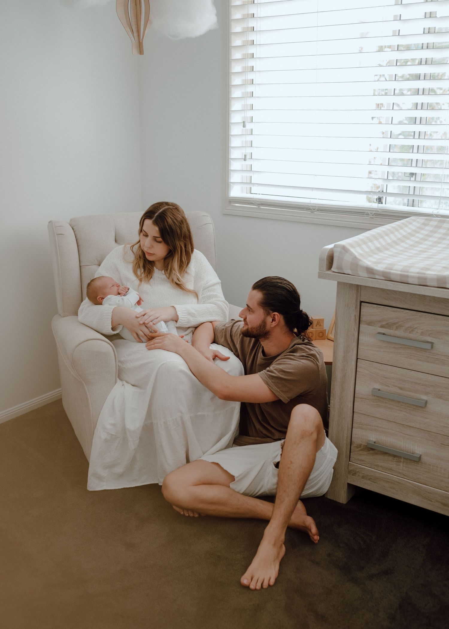 Family relaxing together on white armchair in bright sunlit room with window blinds.