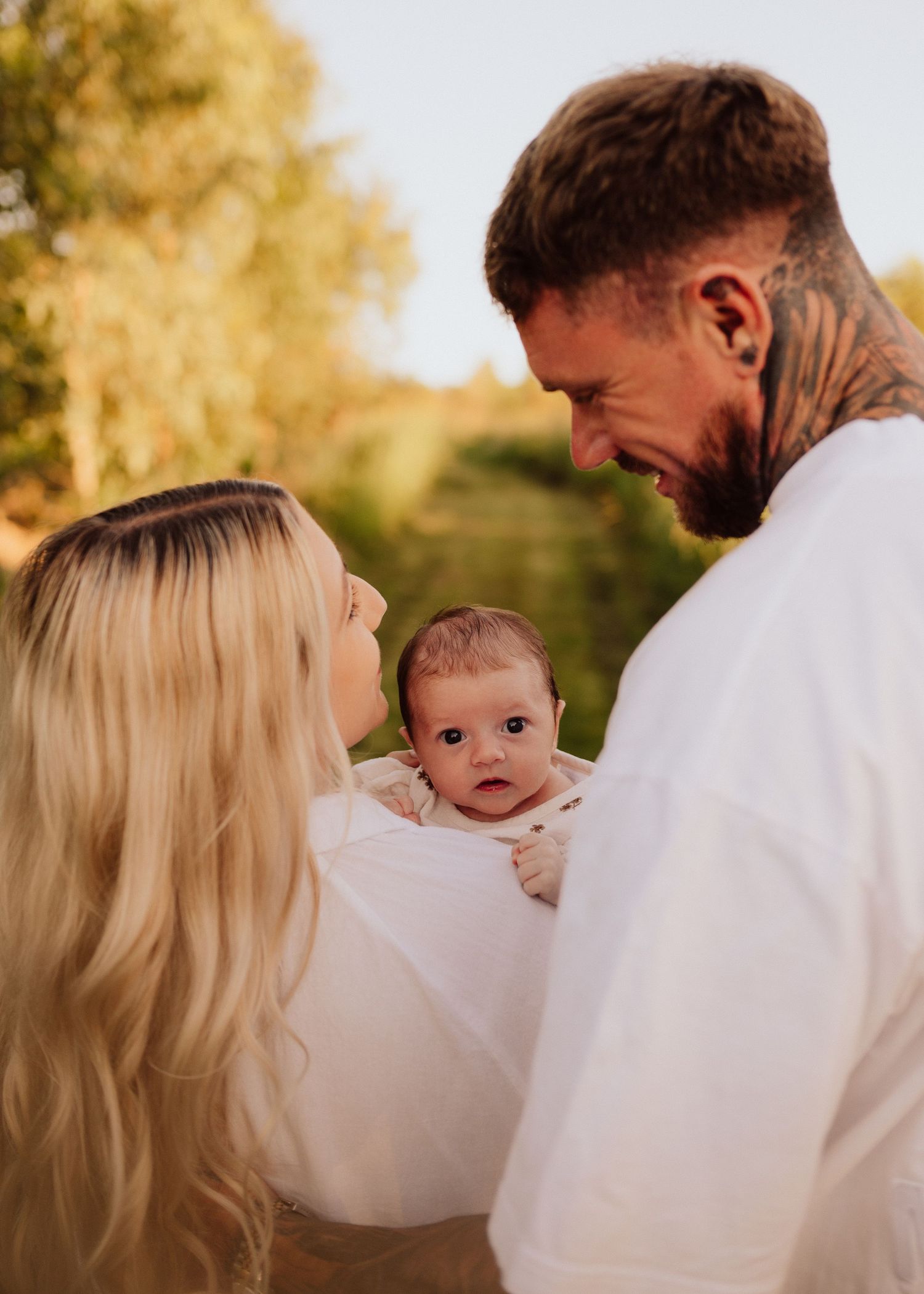 Tattooed father and blonde mother smiling at their newborn baby outdoors in warm golden light.