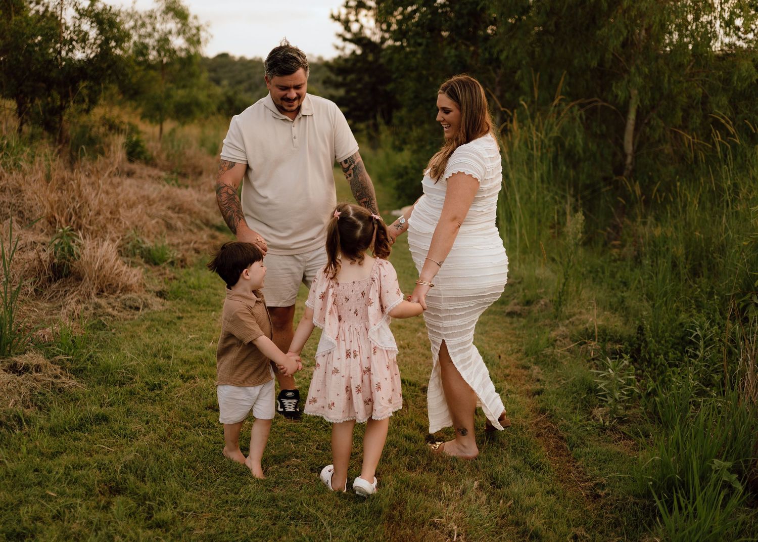 A family walks together on a grassy path during sunset, wearing casual white outfits with a child in a pink dress.