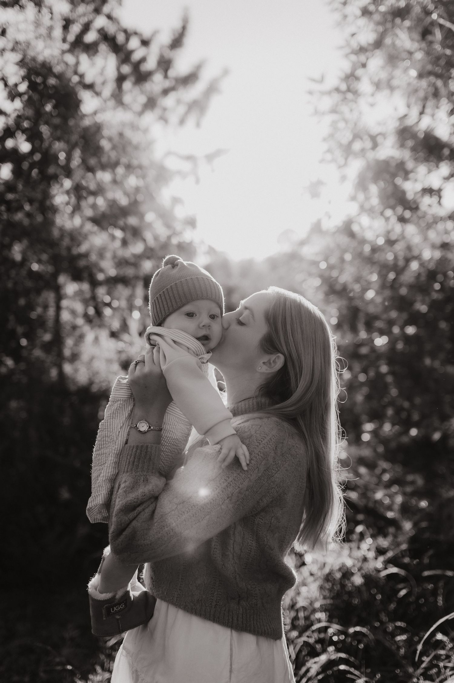 Black and white photo of a mother kissing her baby outdoors, surrounded by trees with soft backlit sunlight.