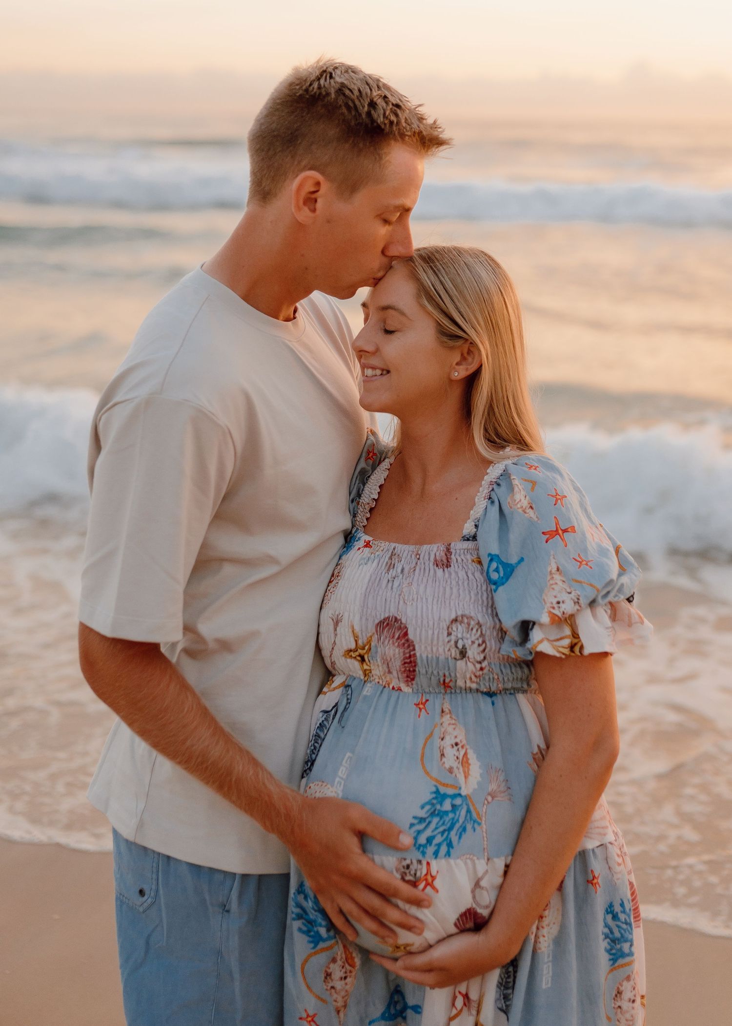 A couple shares tender moments on a beach at sunset wearing casual attire and a floral dress.