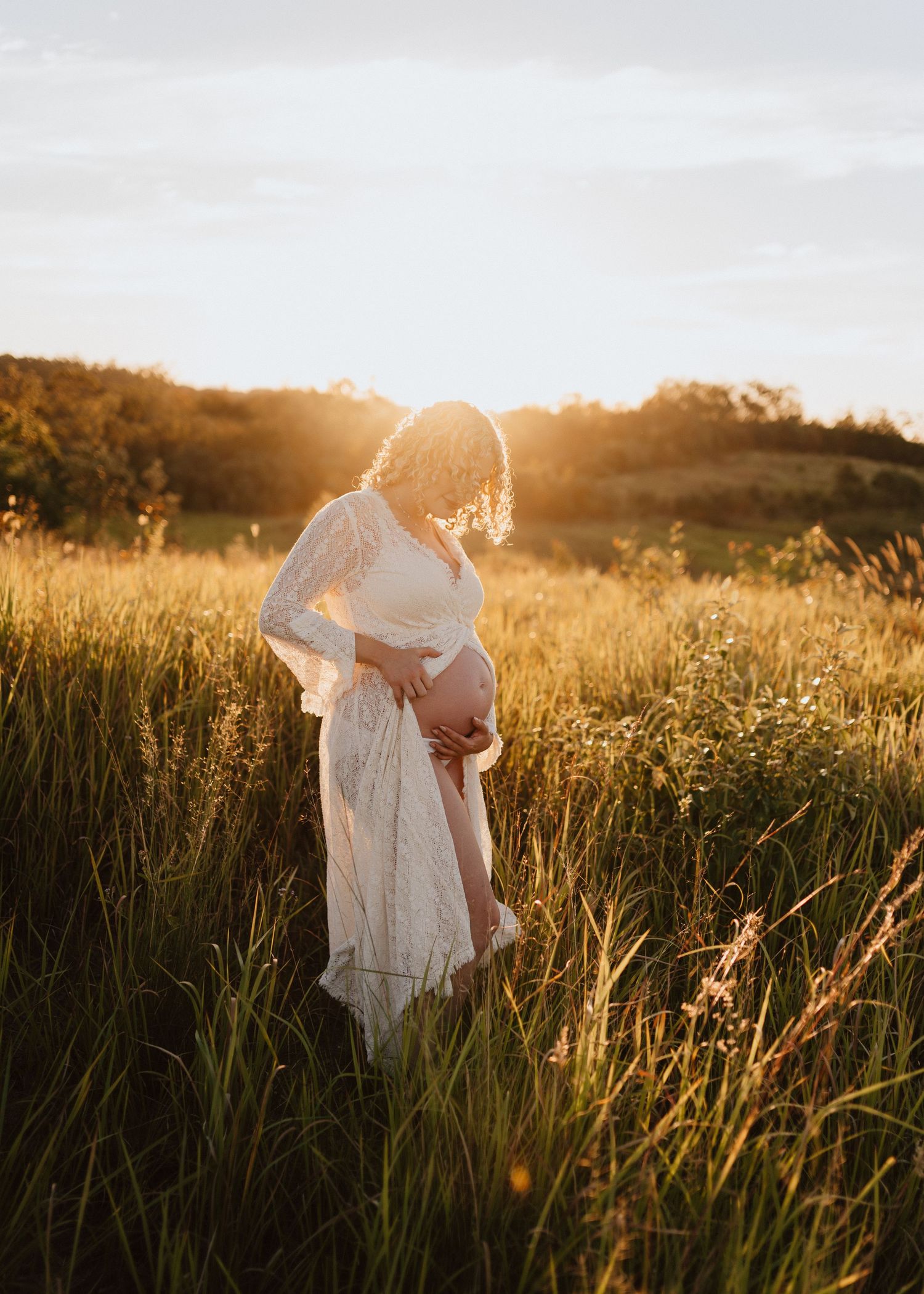 Silhouette of pregnant woman in white dress standing in golden field at sunset with sunlight streaming through tall grass.