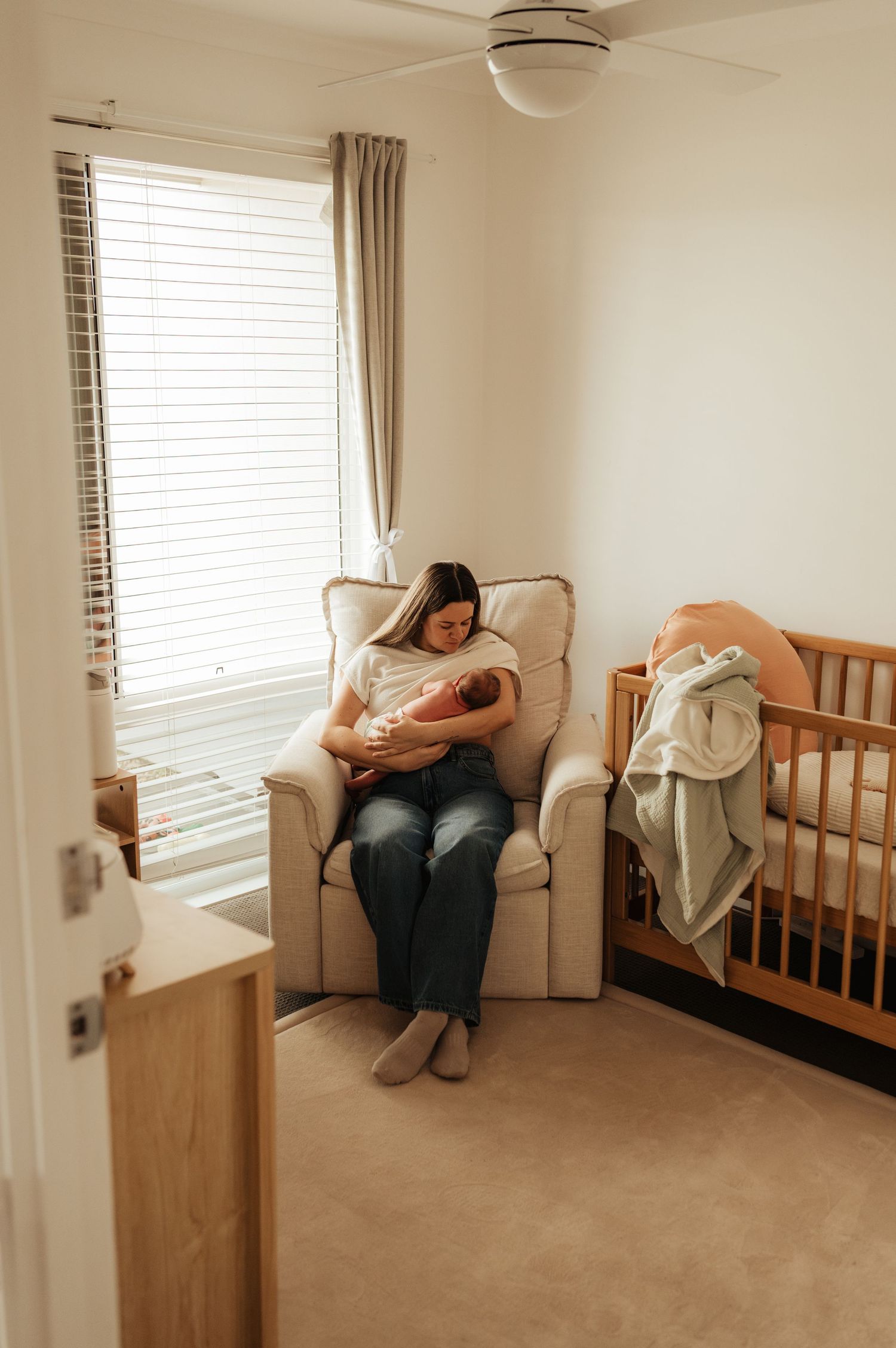Mother cradling newborn baby in nursery glider chair beside wooden crib, bathed in soft natural window light.