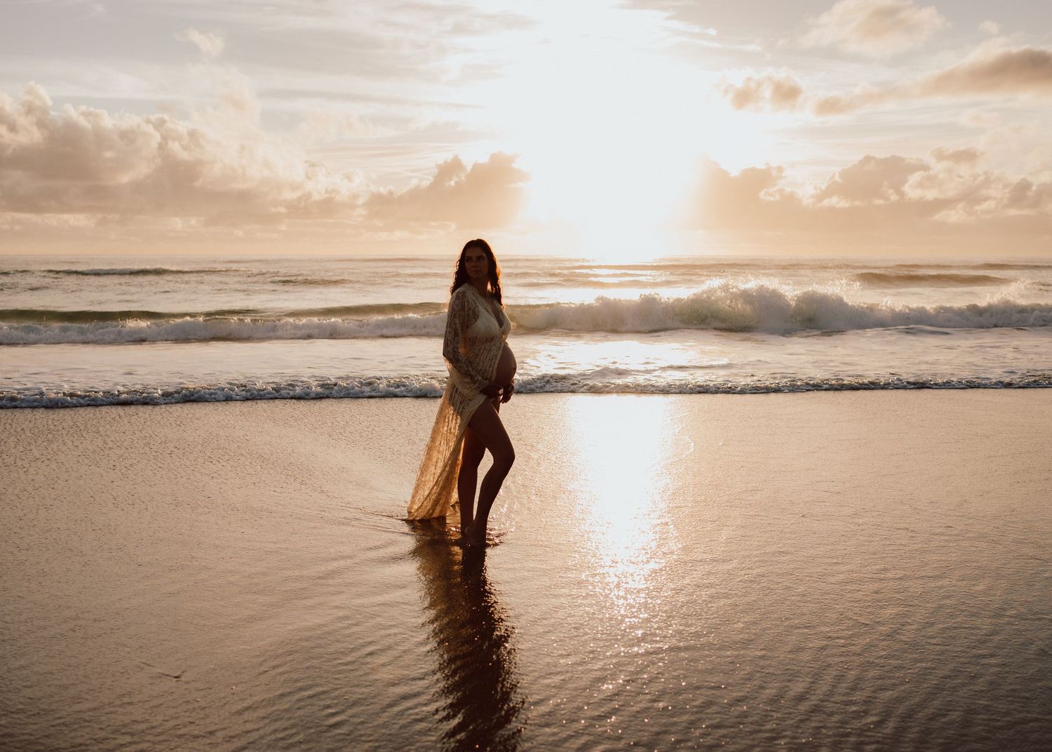 A silhouetted figure in a flowing dress stands at the edge of ocean waves during a golden sunset.