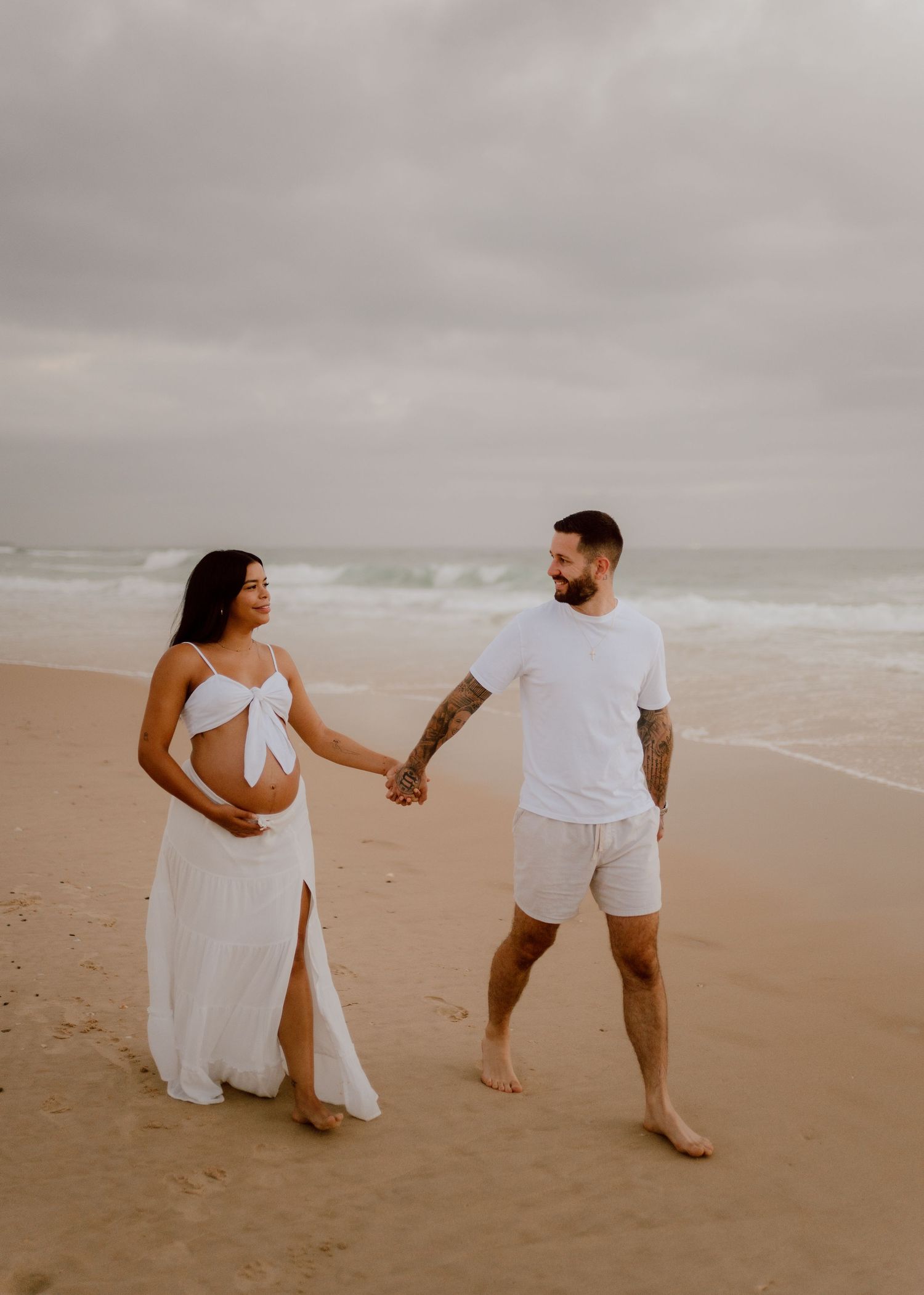 A couple in white outfits holds hands while walking along a misty beach shoreline in a romantic sequence of photos.