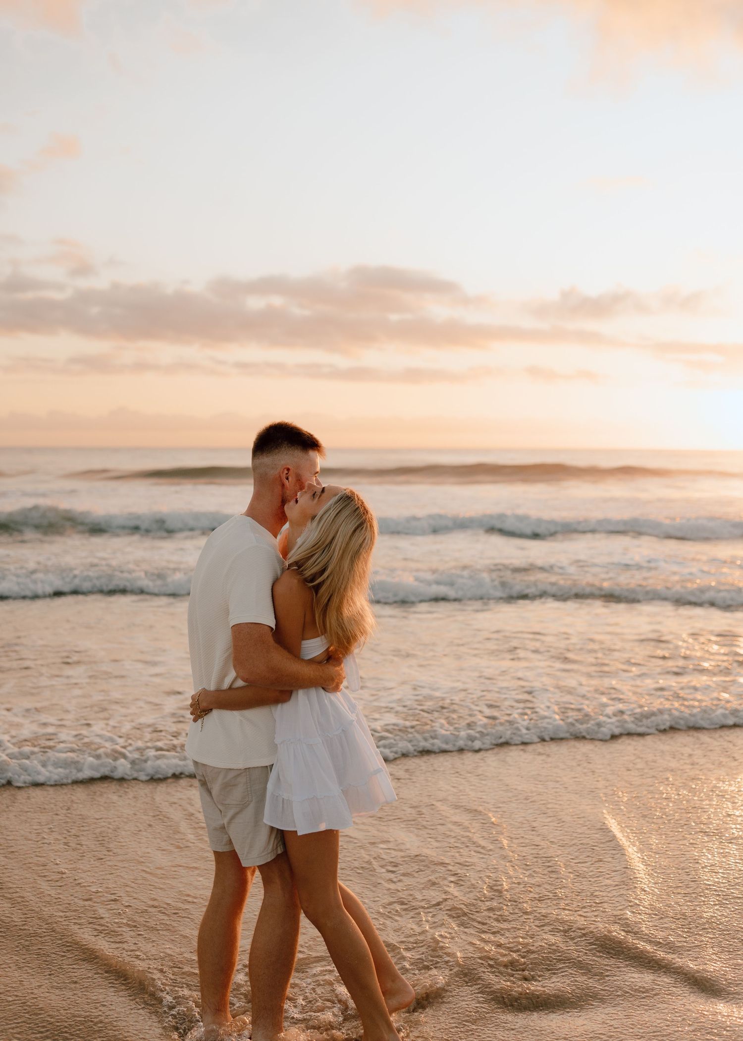 A couple embraces on a sandy beach at sunset with gentle waves rolling in along the shoreline.