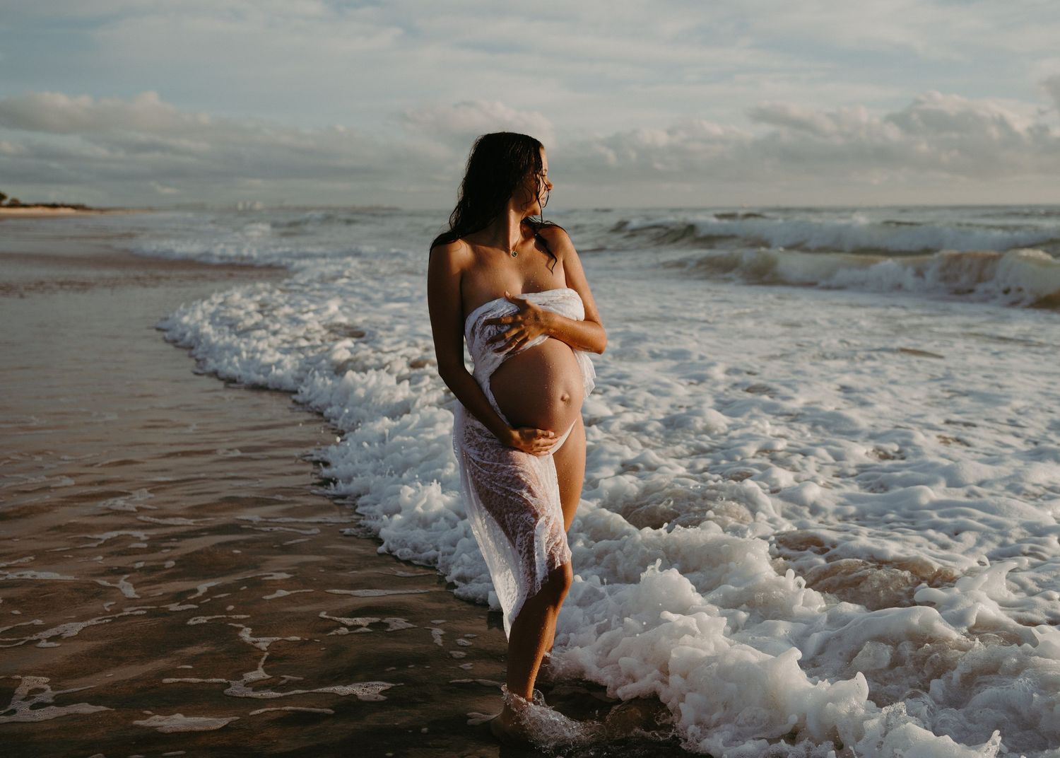 Ocean waves crash around a figure in white as they dance along the shoreline at dusk.