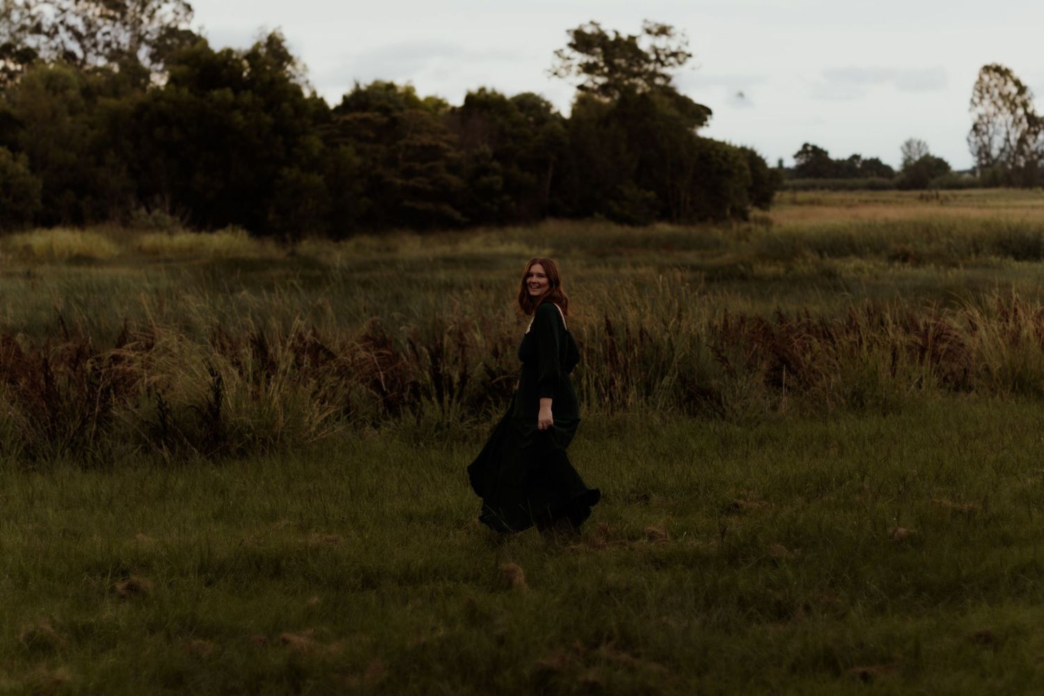 A figure in a flowing black dress stands alone in a moody autumn field at dusk.