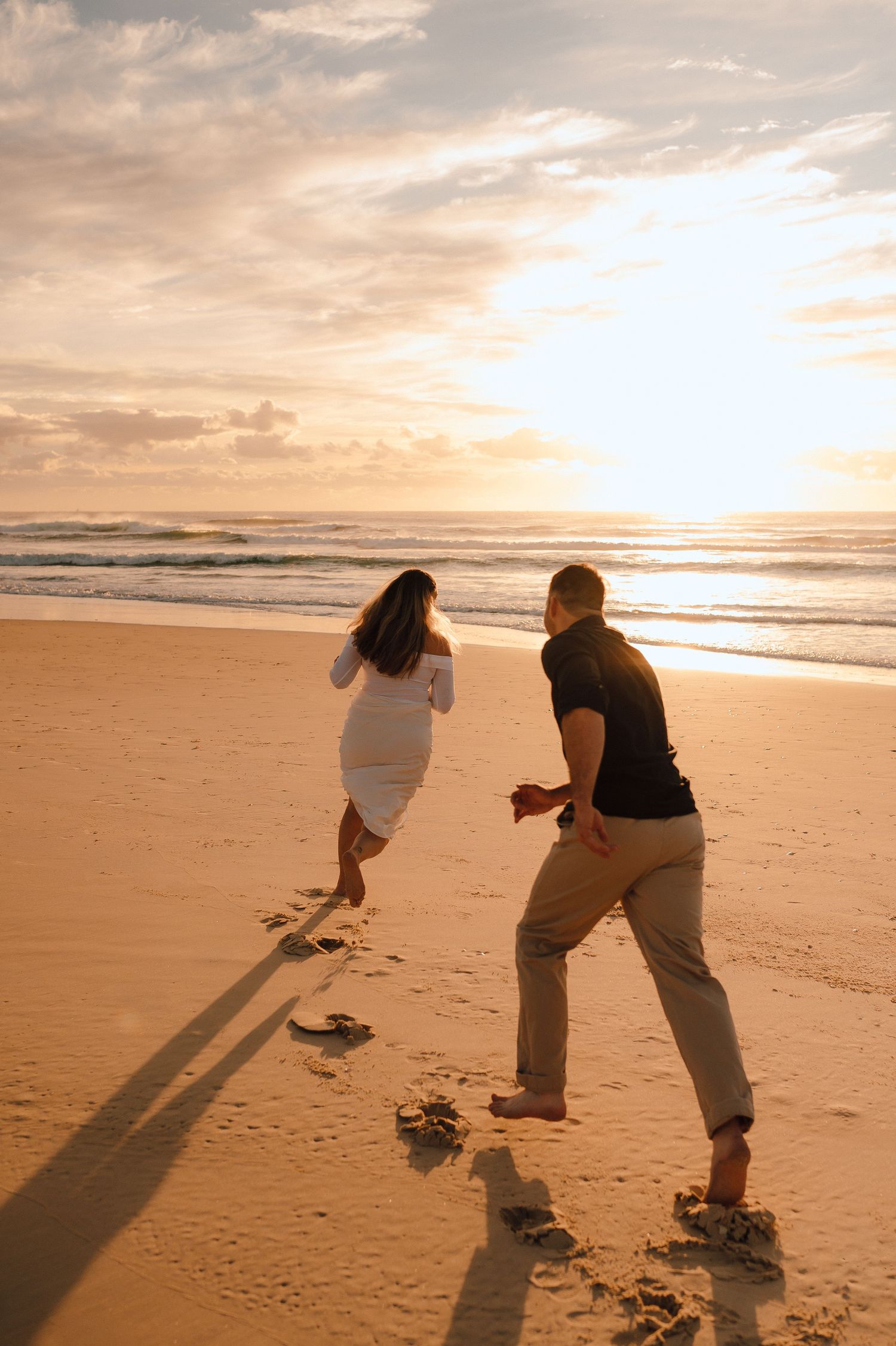 Couple running barefoot on sandy beach at golden sunset, leaving footprints in the sand near ocean waves.