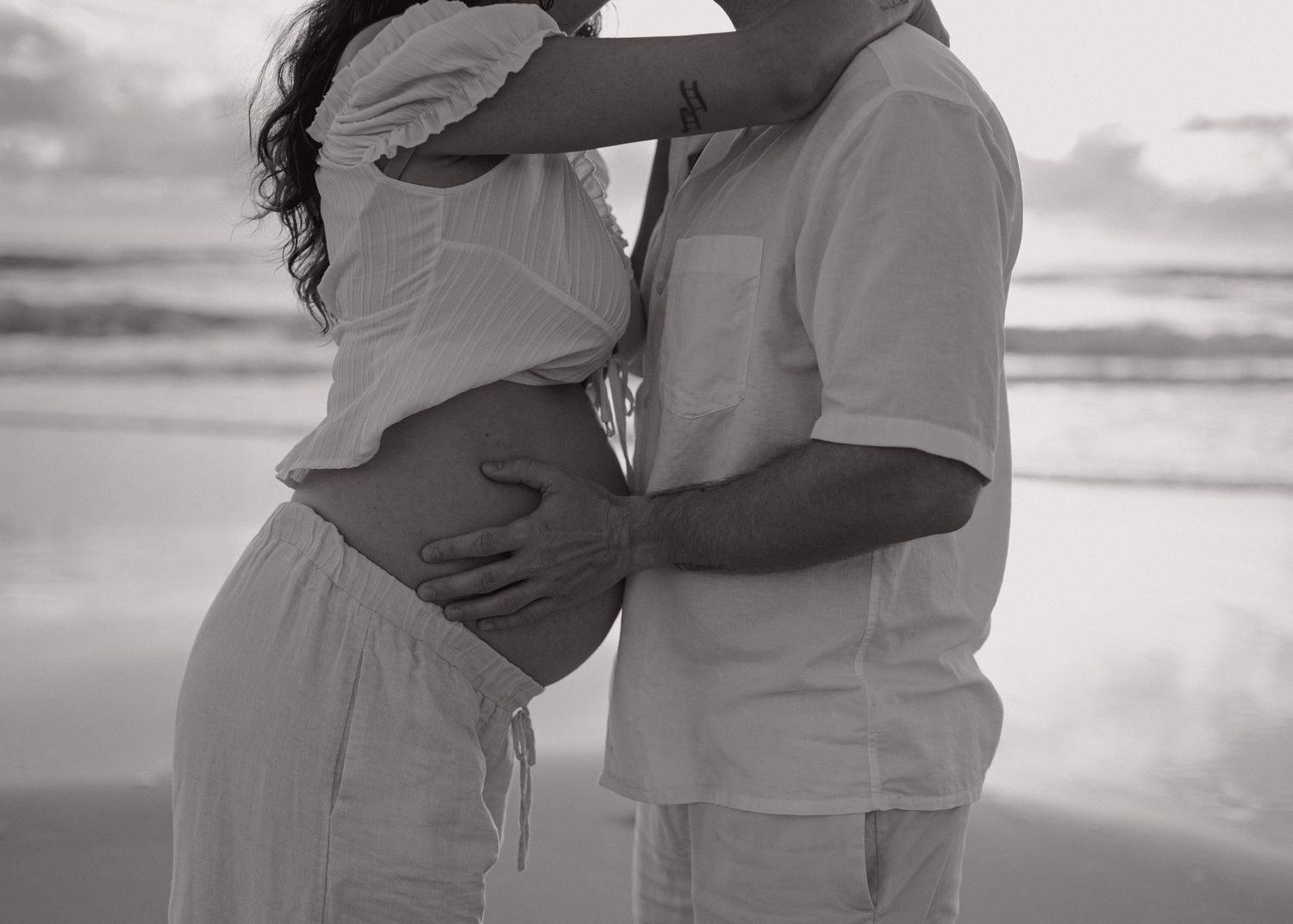 A tender black and white beach scene of a couple embracing by the ocean at sunset.