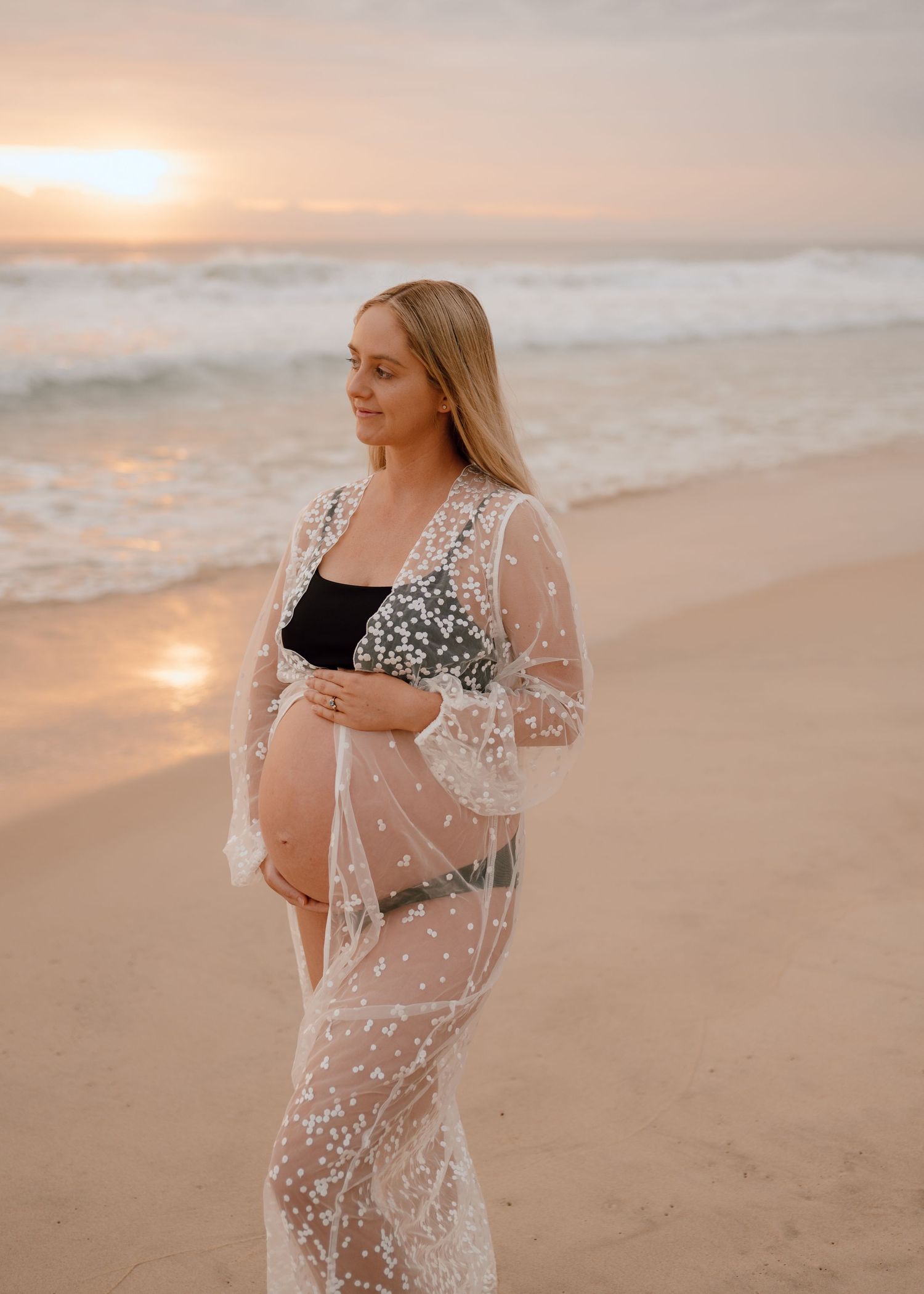 Expectant mother in sheer lace coverup poses on beach at sunset with waves and golden light in background.