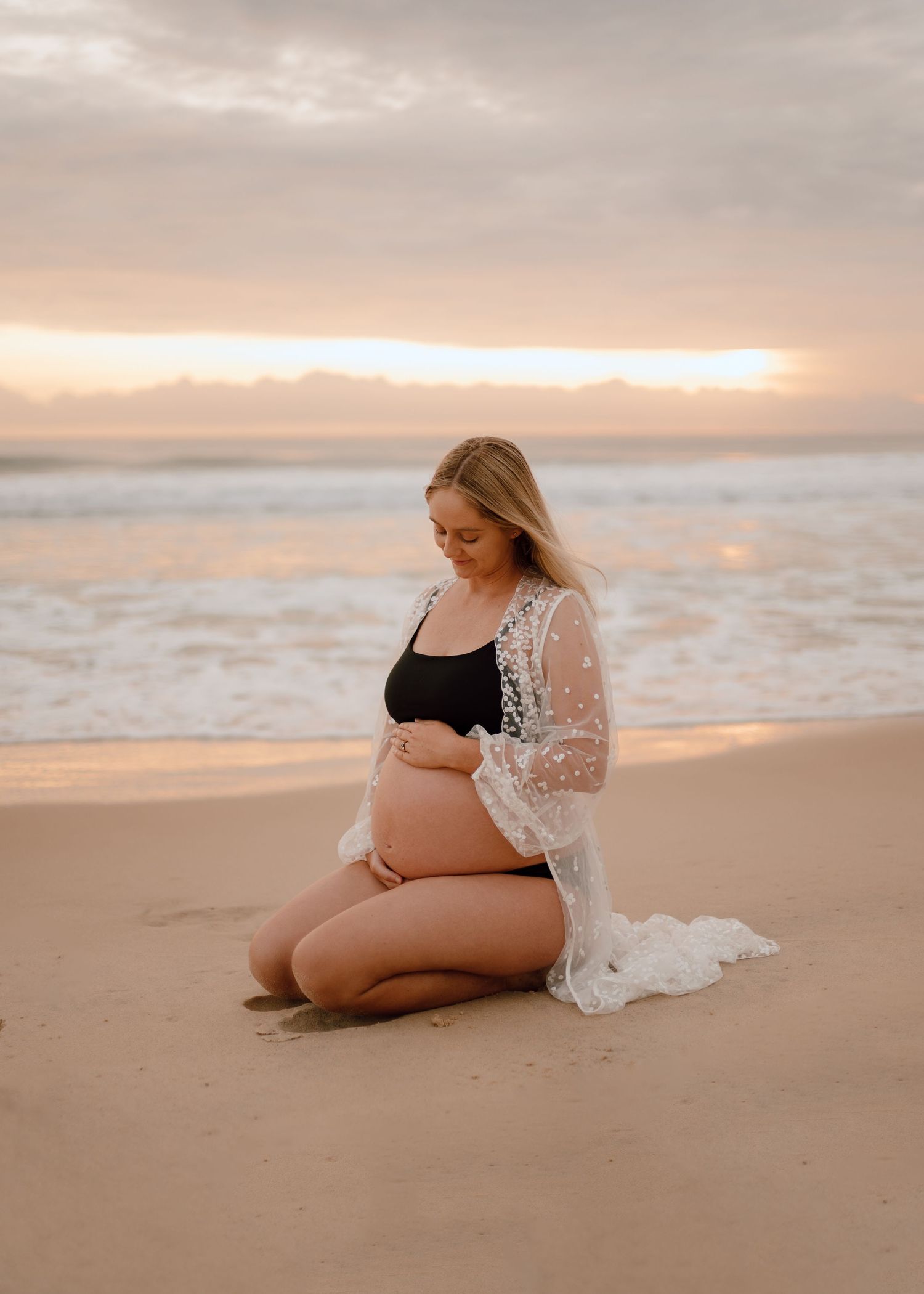 A maternity photoshoot on a sandy beach at sunset with soft pink and orange sky in the background.