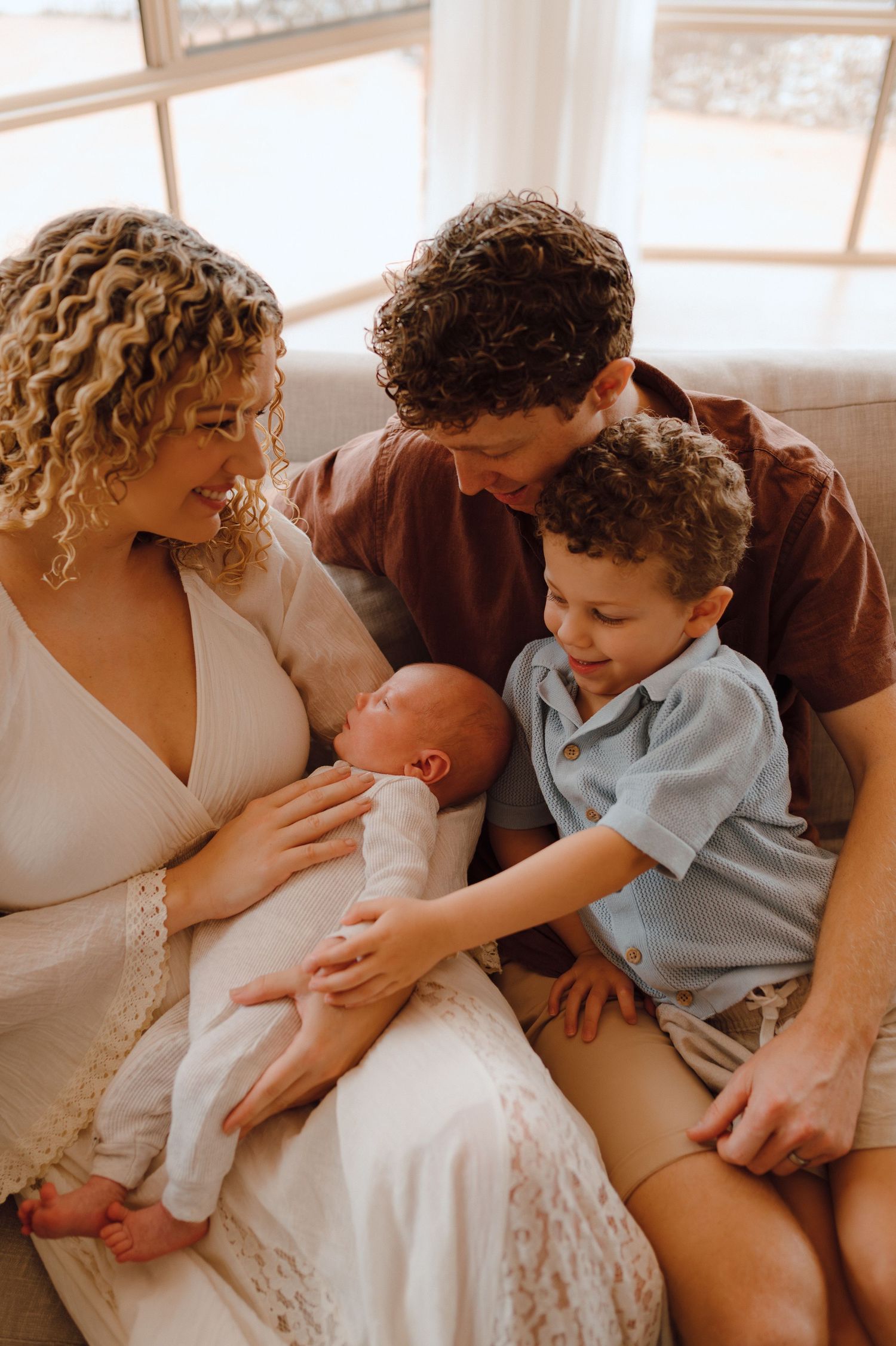 A family of four admires their newborn baby on a couch, with curly-haired parents and an older child gently touching the infant.