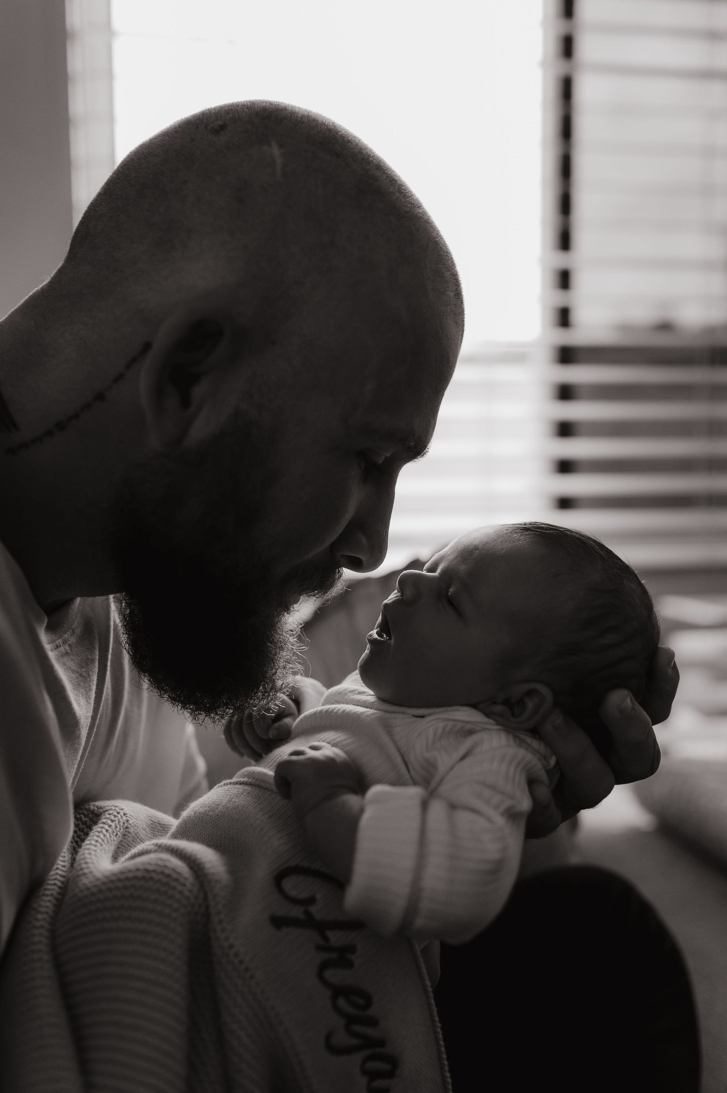 Black and white photo of a bald bearded man tenderly holding and gazing at his newborn baby wearing a onesie.