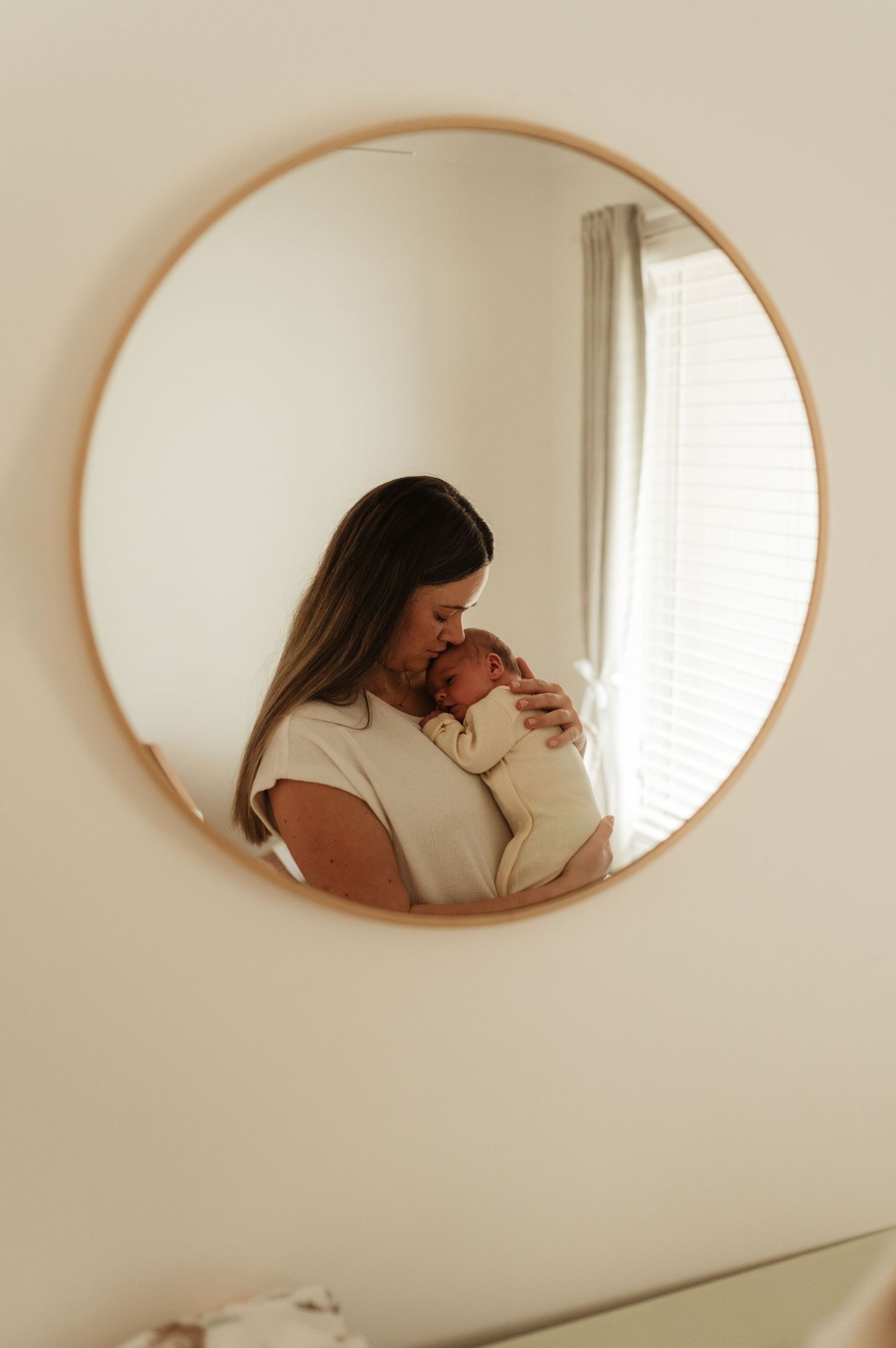 A mother tenderly holds her newborn baby, reflected in a round wood-framed mirror on a soft white wall.