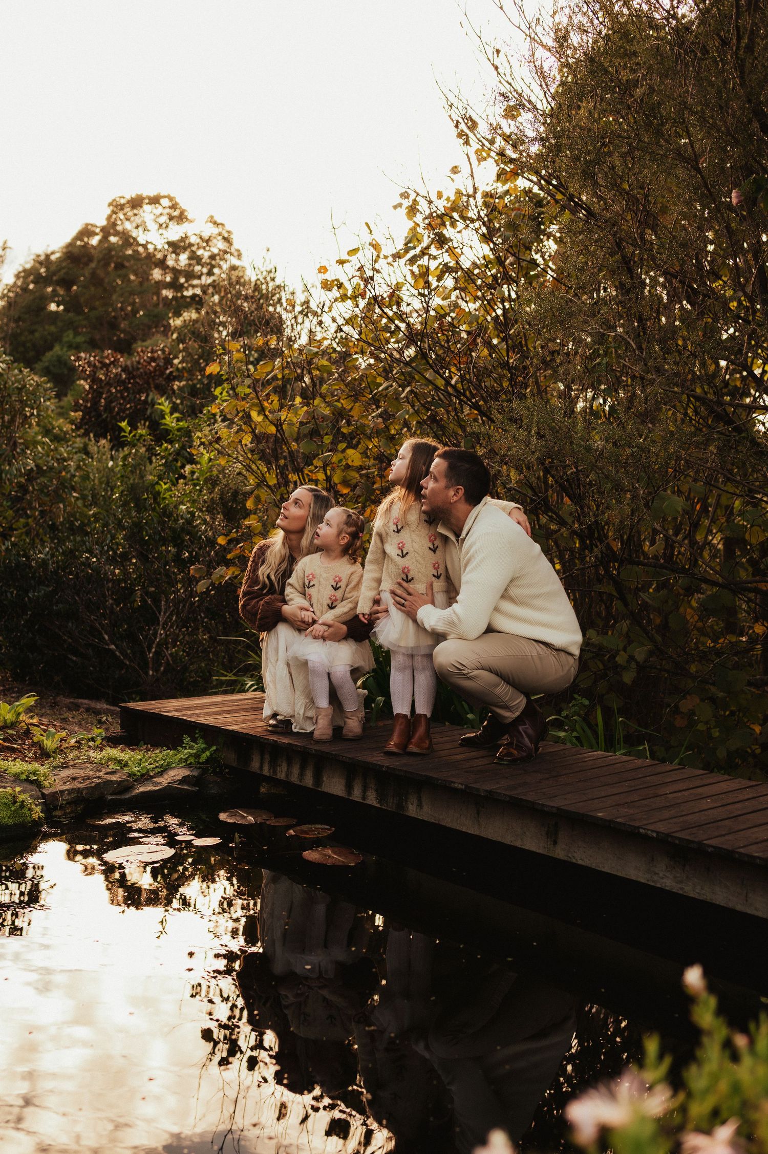 Family of four posing on a wooden bridge over a pond surrounded by lush greenery during golden hour.